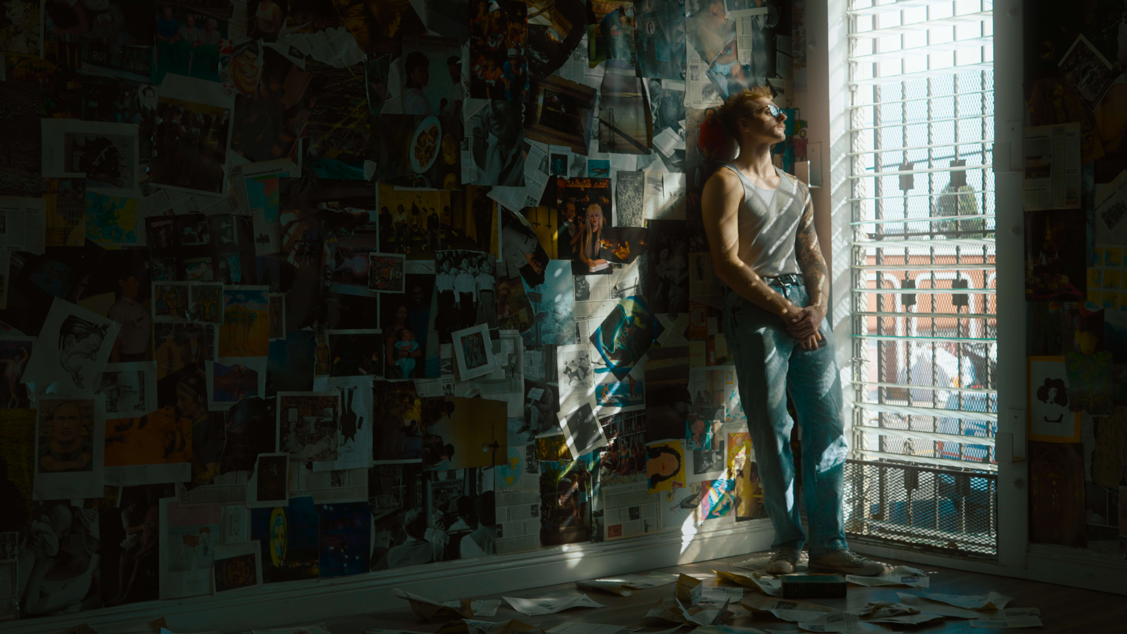 A person stands beside a partially open window in a room filled with photos and papers taped to the walls.