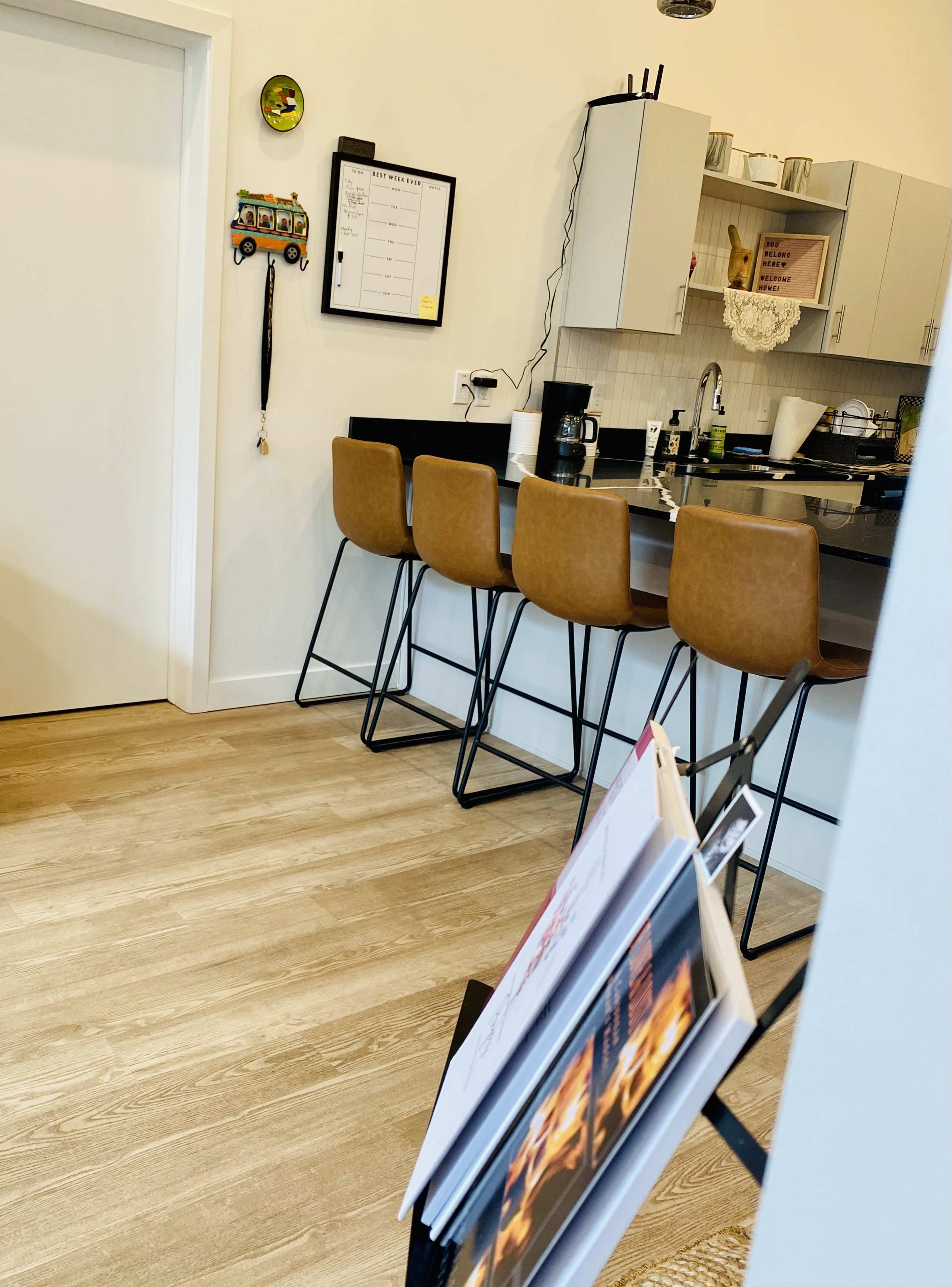 A modern kitchen with a bar counter and four stools, a wall-mounted calendar, and a magazine rack in the foreground.