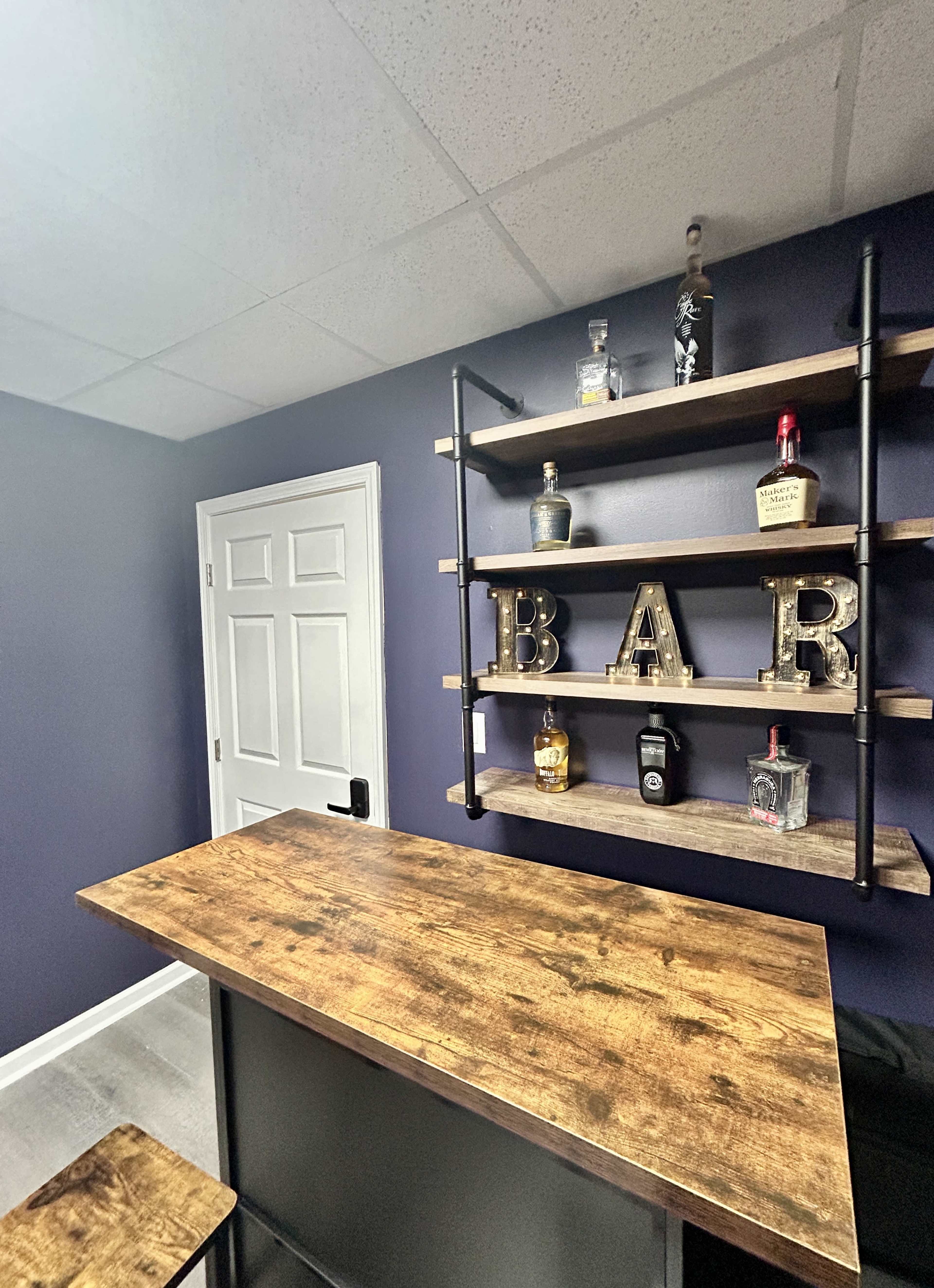 The image shows a small home bar with a wooden countertop, shelves displaying various liquor bottles, and the letters "BAR" in decorative style against a dark purple wall.