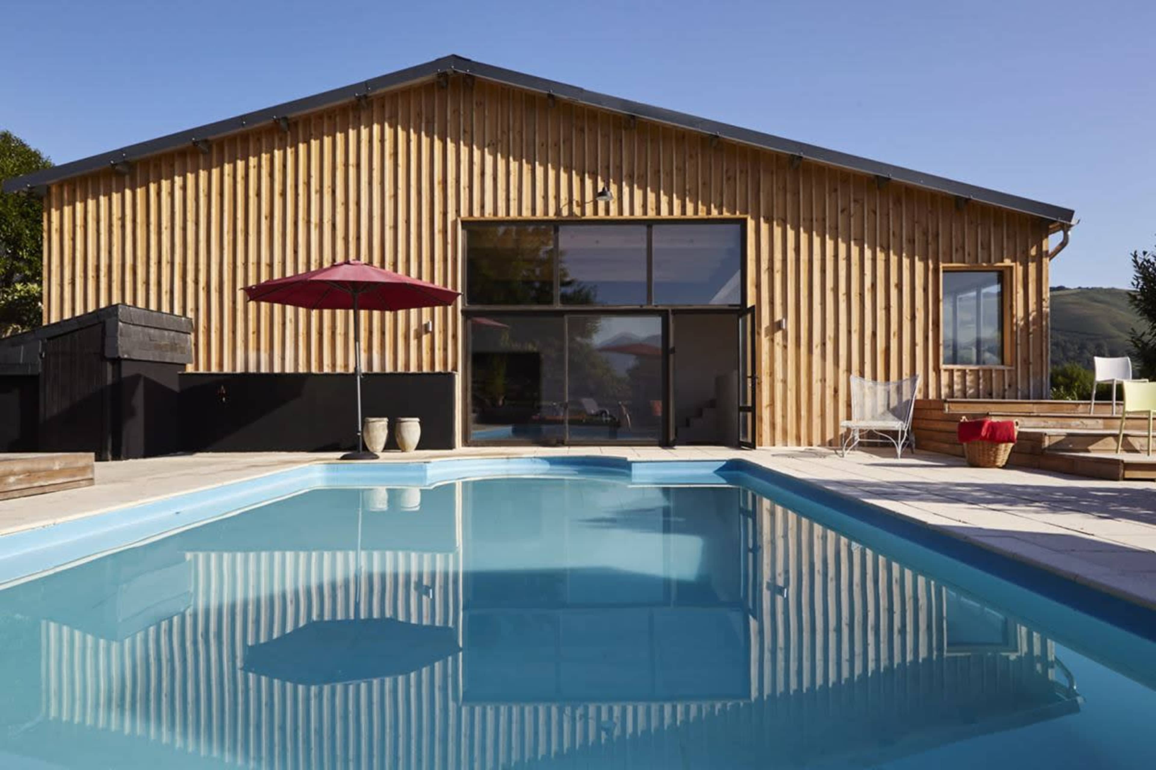 A modern wooden house with large windows overlooks a swimming pool surrounded by a patio.