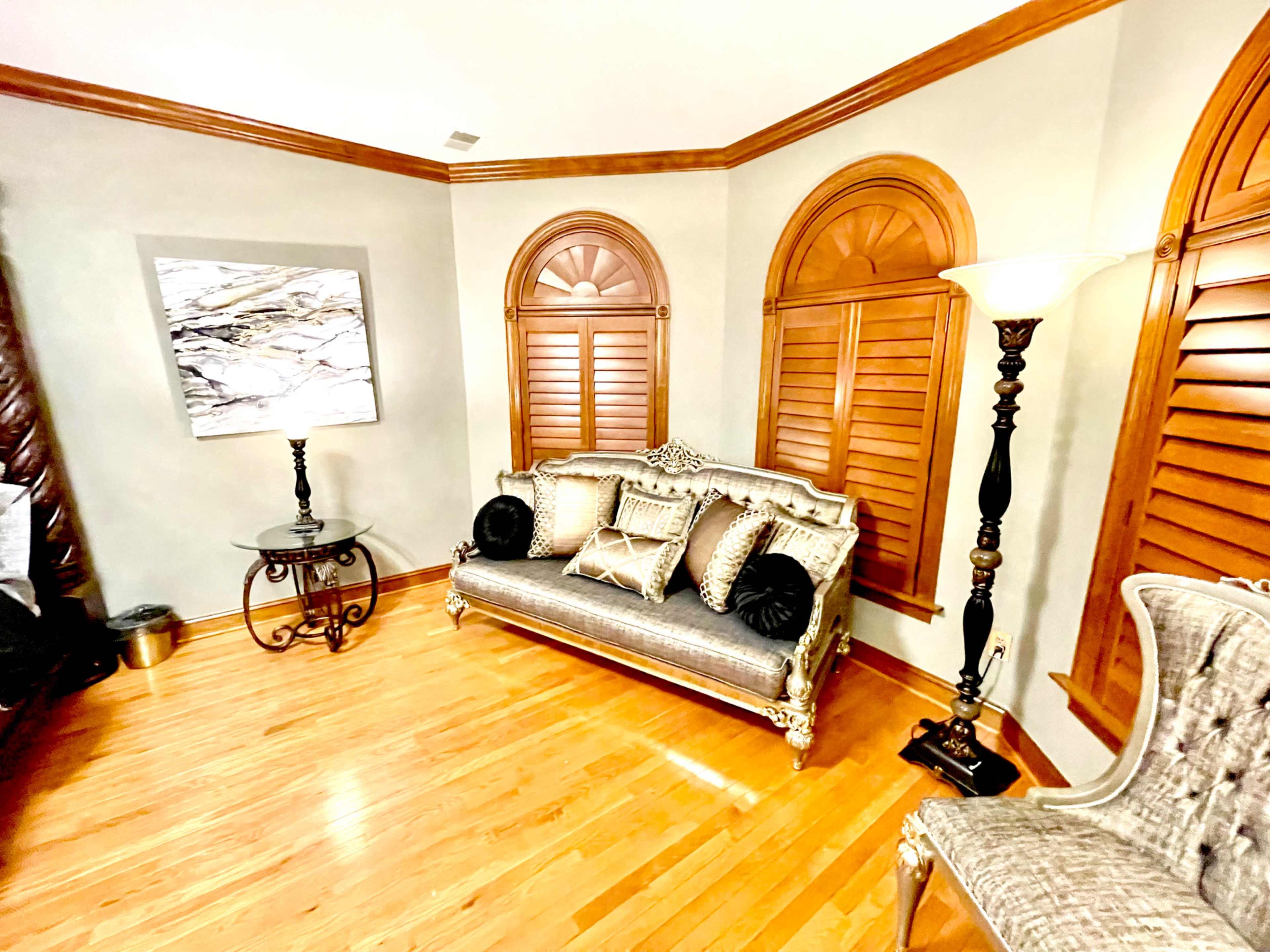 The image shows a living room featuring a decorative couch with pillows, wooden shutters on the windows, and a small side table with a lamp.