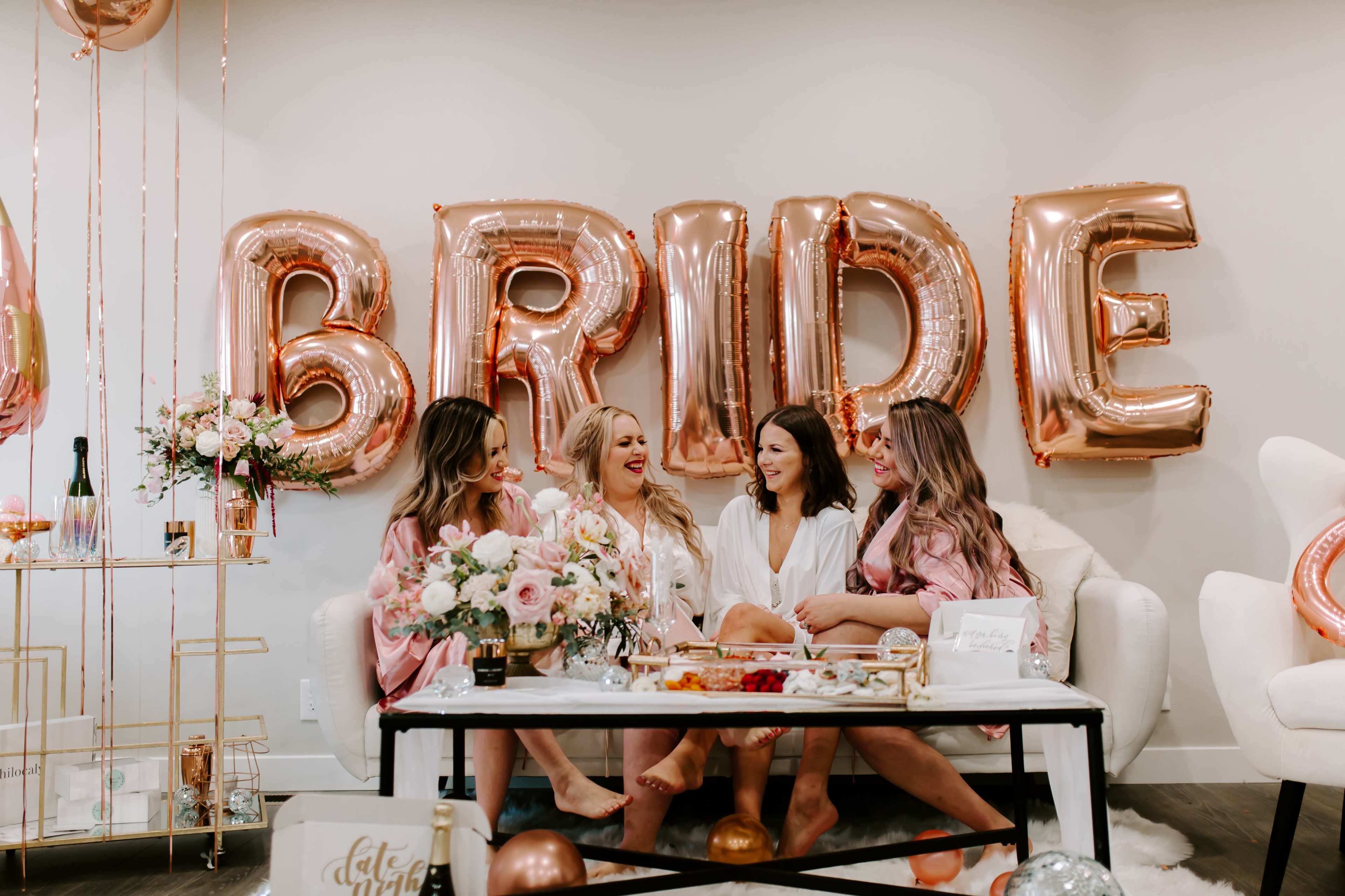 Four women in matching robes celebrate a bridal event, sitting around a table adorned with flowers and snacks, with large rose gold "BRIDE" balloons in the background.