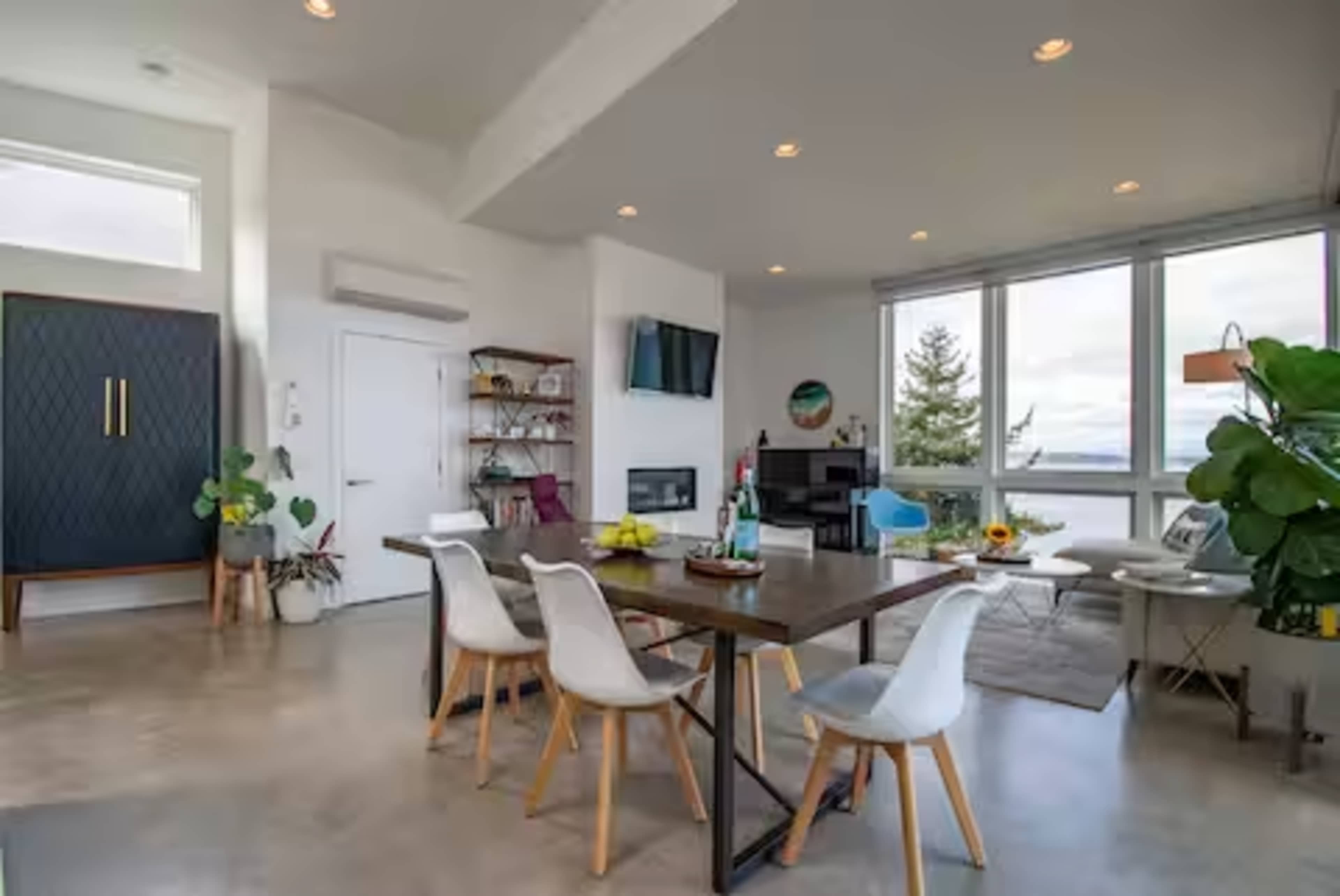 The image shows a modern dining area with a large table and chairs, surrounded by a living space that includes a television, plants, and large windows showcasing a view.