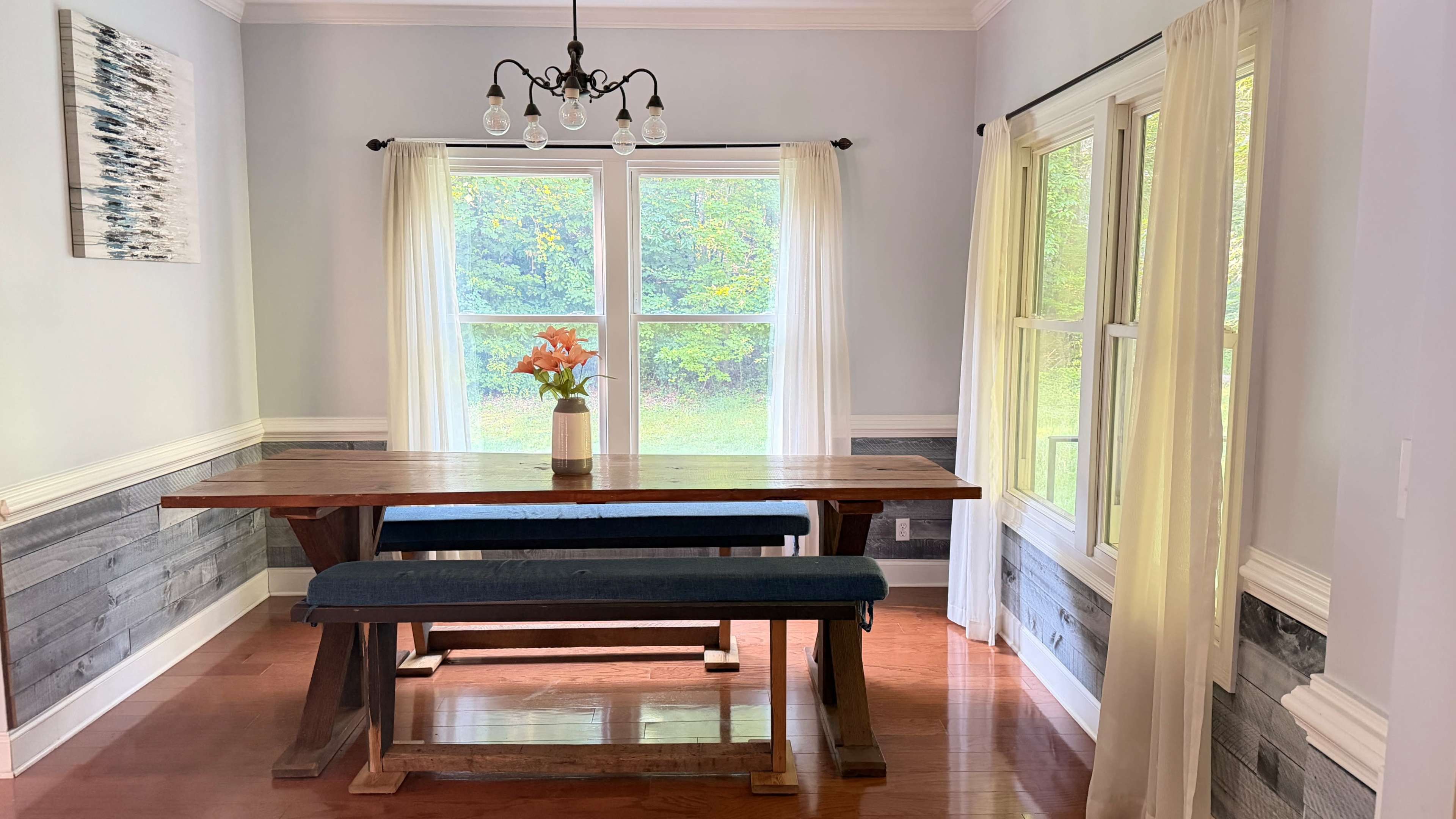 A wooden dining table with a bench is positioned under a chandelier in a bright, airy room with large windows and decorative curtains.
