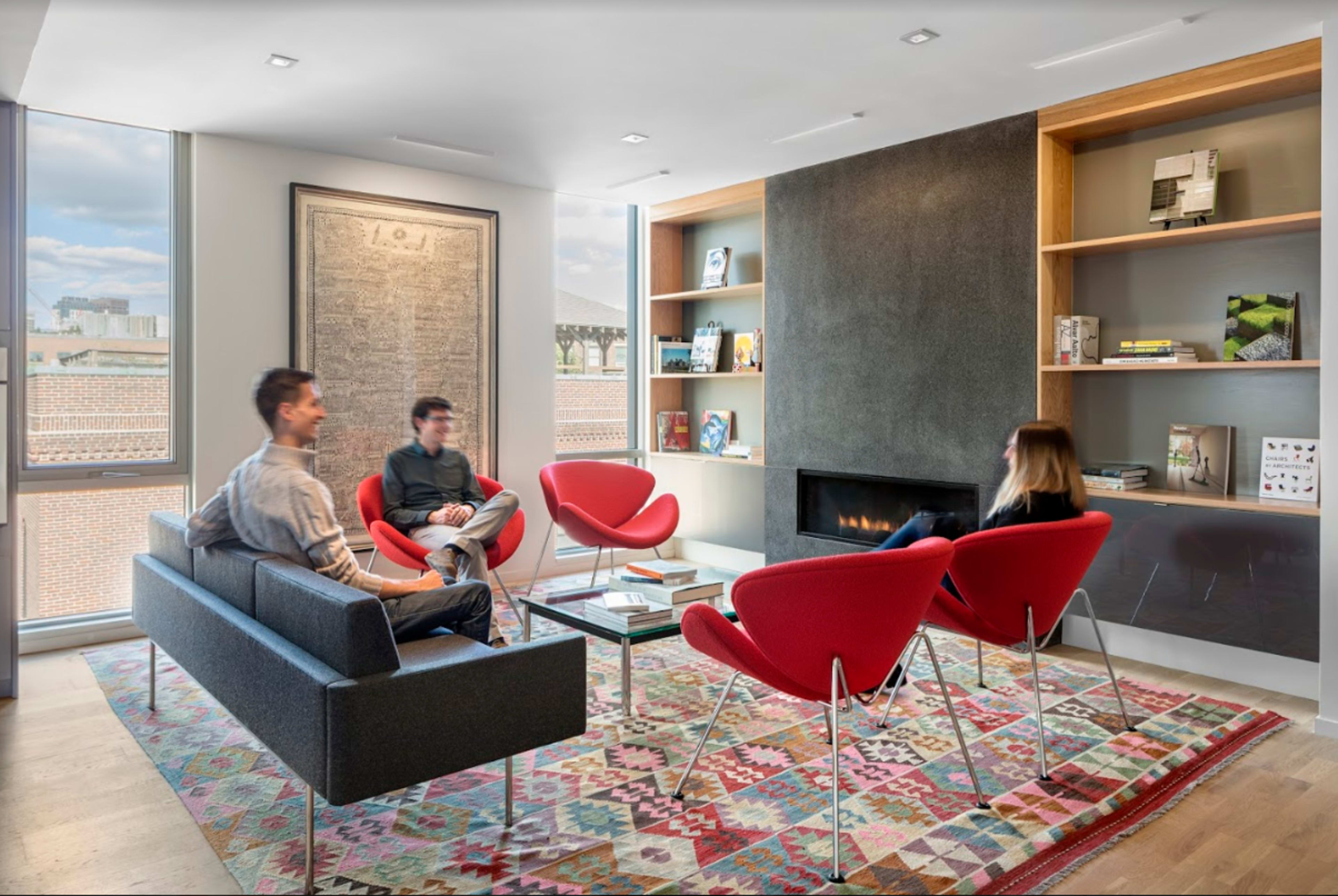 A modern living room features four people seated in red chairs and a gray sofa, with a fireplace and bookshelves in the background.