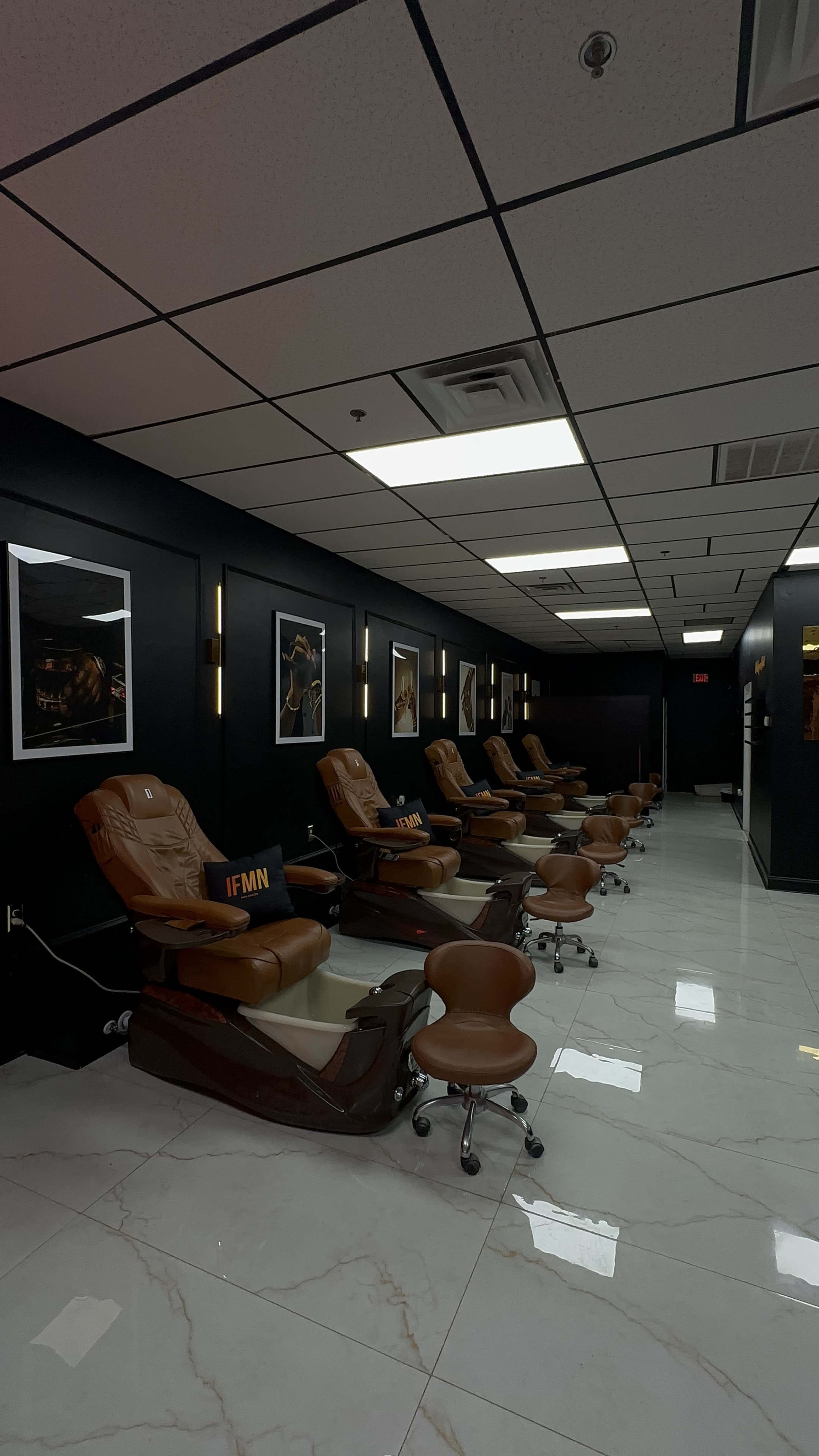 The image shows a modern nail salon with several brown pedicure chairs lined up against a black wall adorned with framed pictures.
