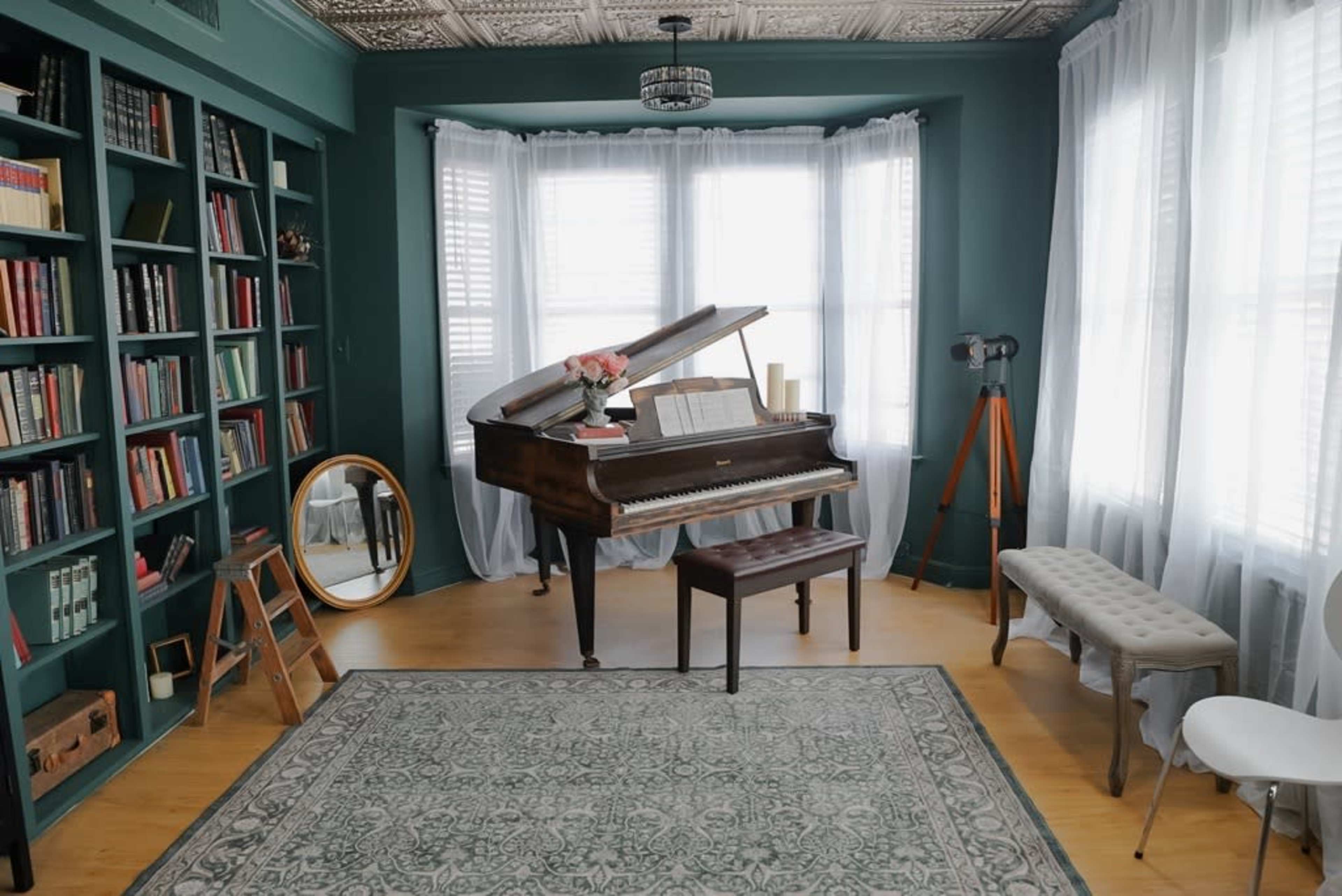 The image shows a cozy room with a grand piano, a wooden bench, a large mirror, and bookshelves filled with books, all under a decorative ceiling.