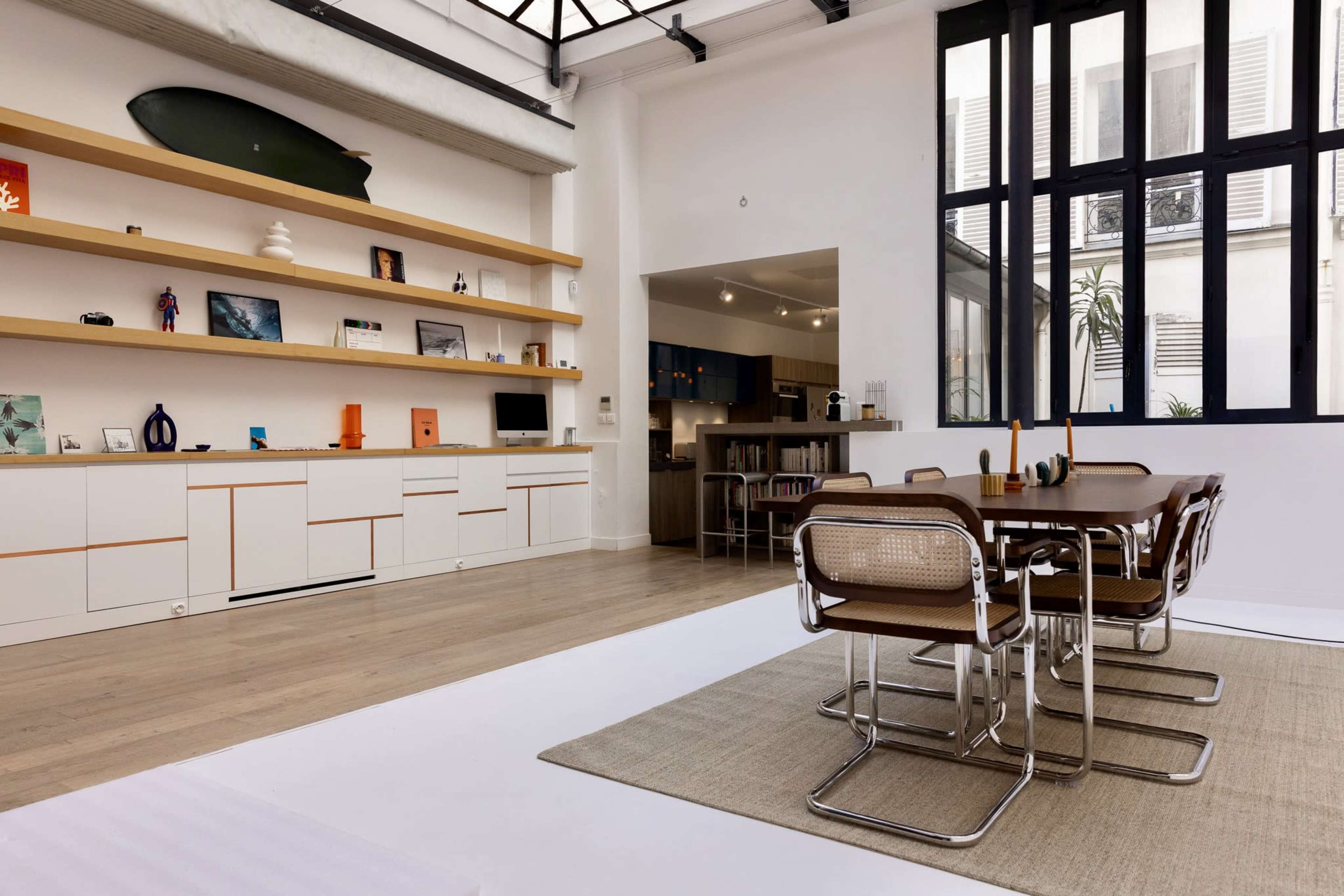 The image depicts a modern, open-concept living space featuring a dining area with a table and chairs, a sleek shelving unit, and an adjacent kitchen area.