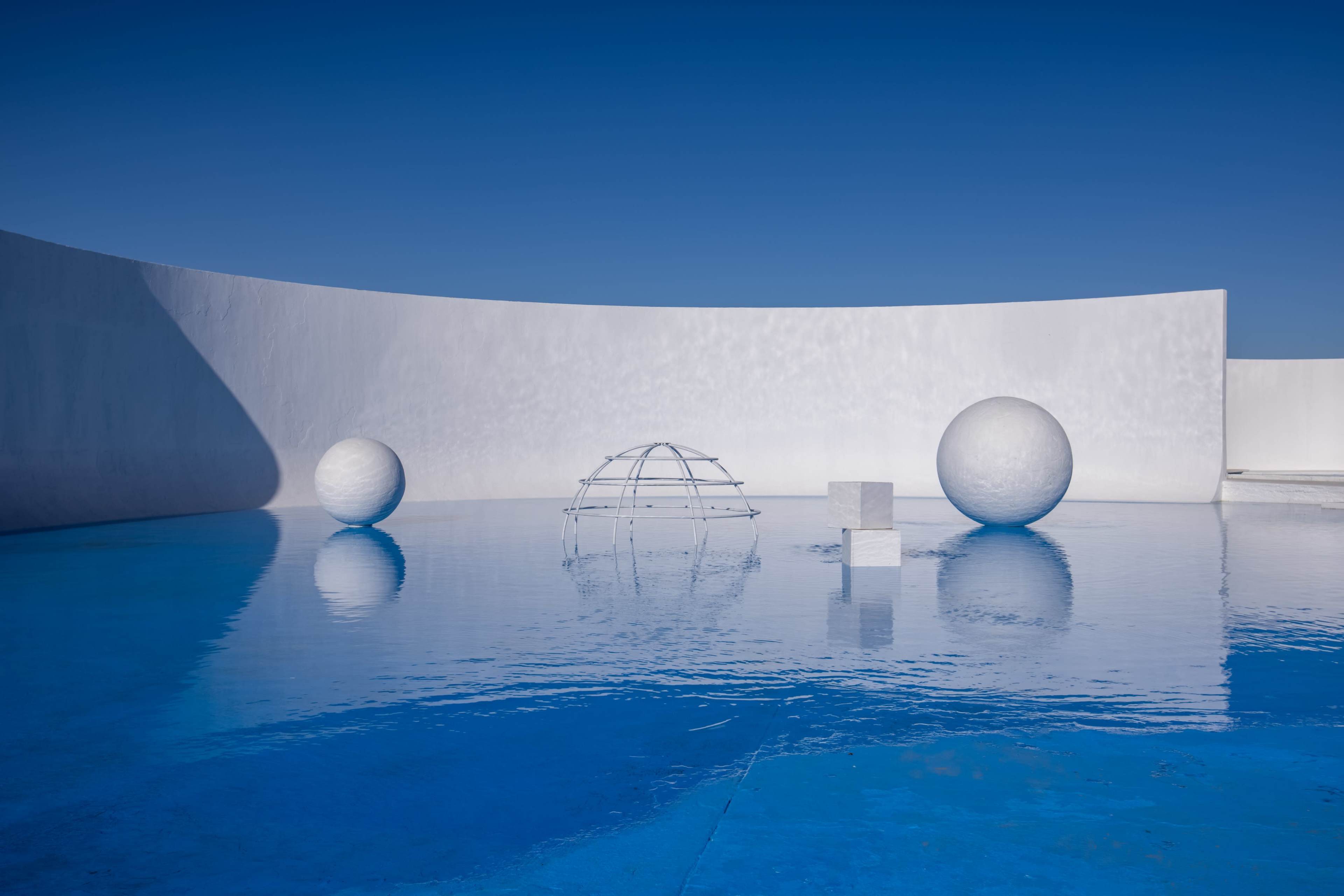 The scene features a minimalist outdoor space with large white geometric sculptures surrounded by a tranquil blue pool of water under a clear sky.