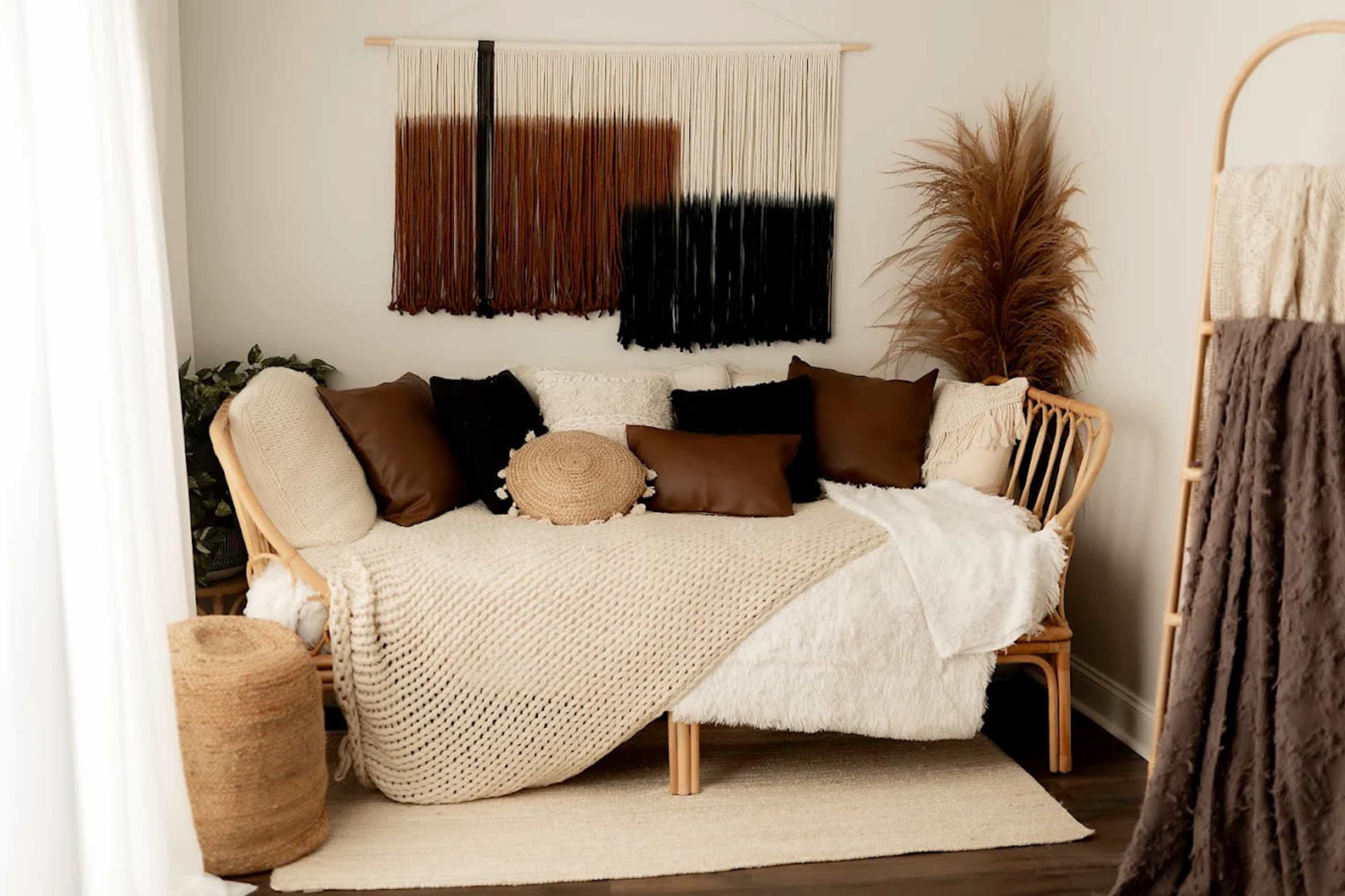 The image features a cozy, styled living space with a woven couch adorned with various decorative pillows, a textured throw blanket, and a wall hanging made of yarn in earthy tones.