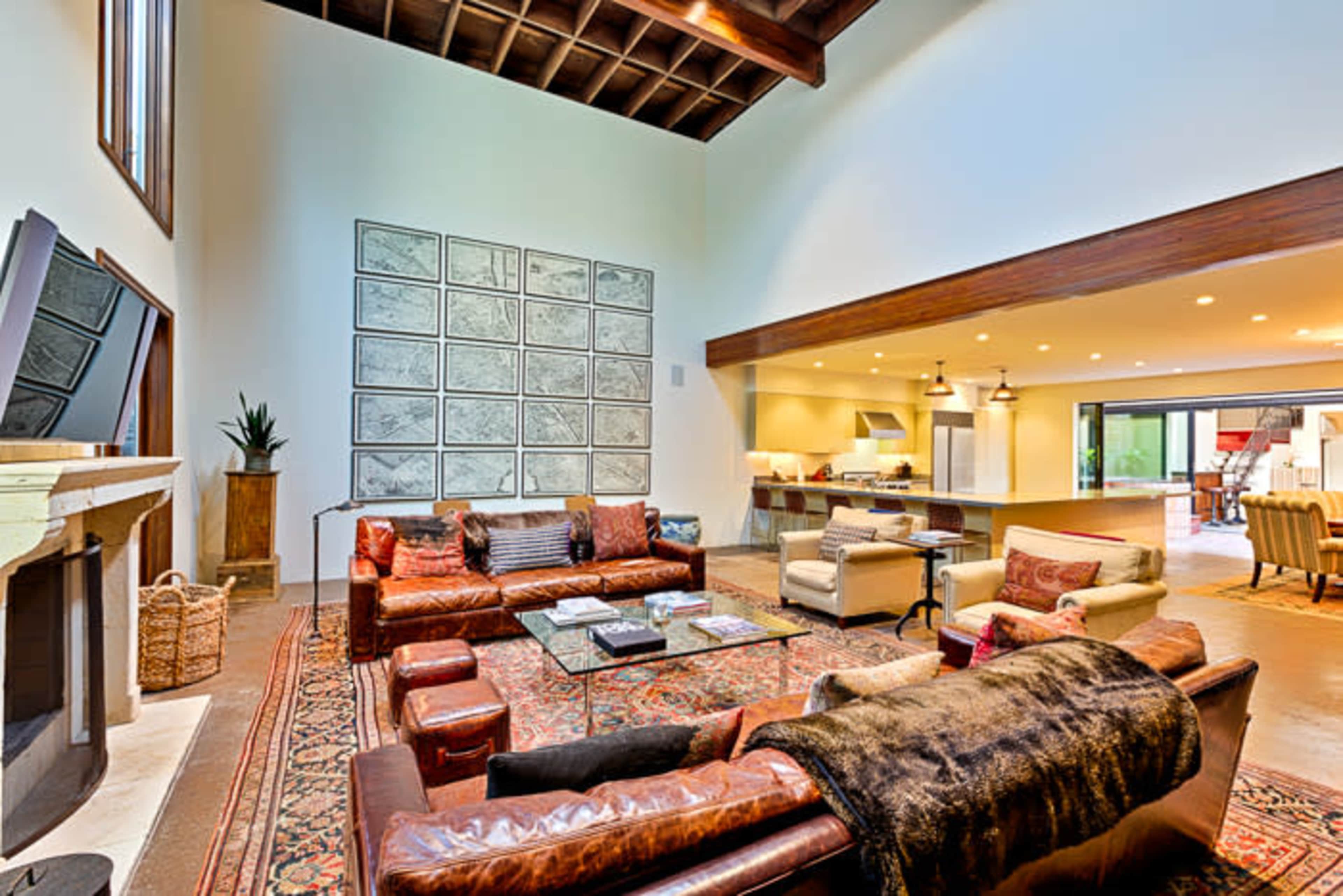The image shows a spacious living room with high ceilings, featuring a large leather sofa, a coffee table, and a series of framed artworks on the wall, along with an open view of a kitchen area in the background.