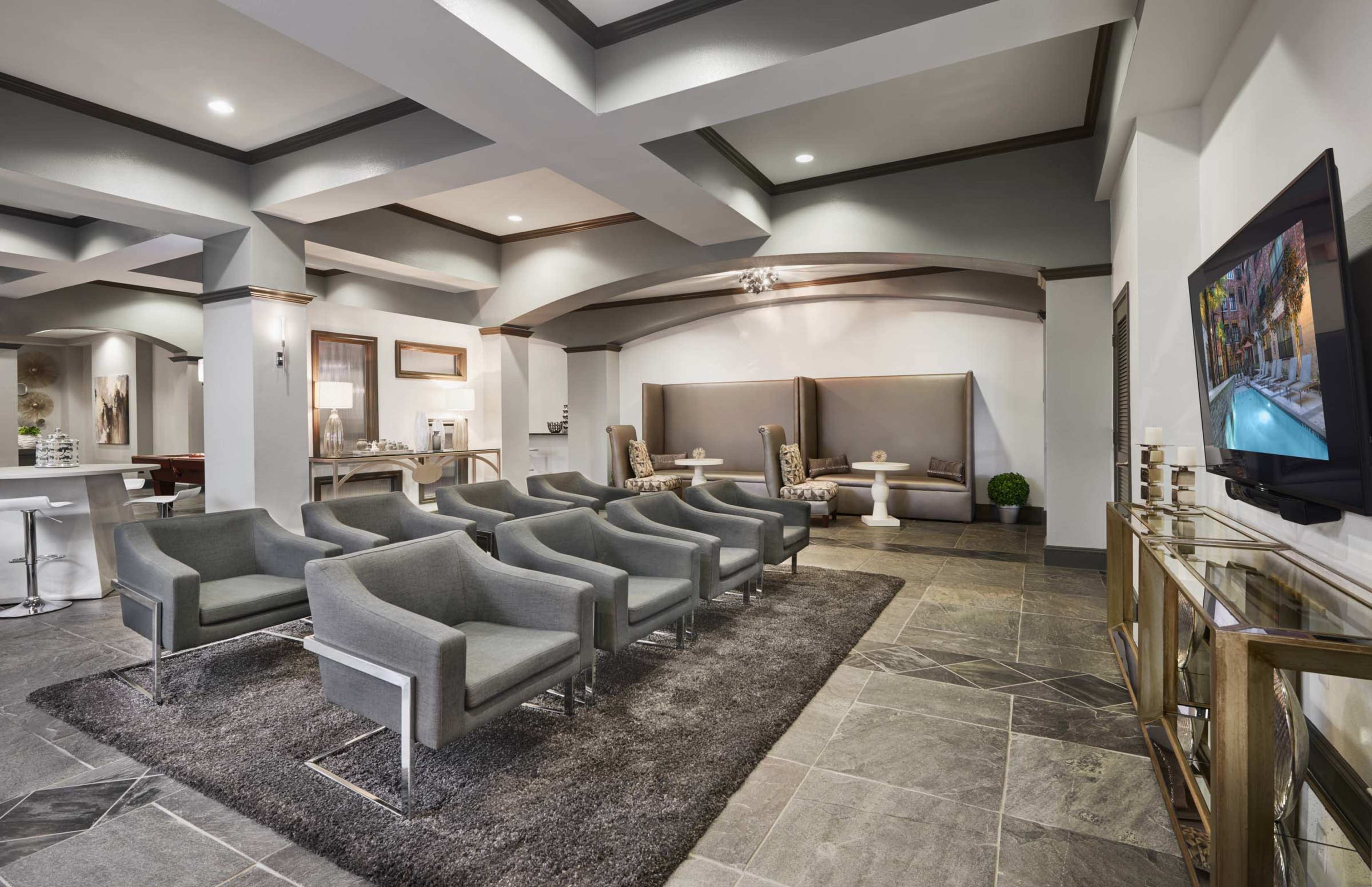 A modern waiting area featuring gray chairs arranged on a large rug, with a small seating nook and a television mounted on the wall.