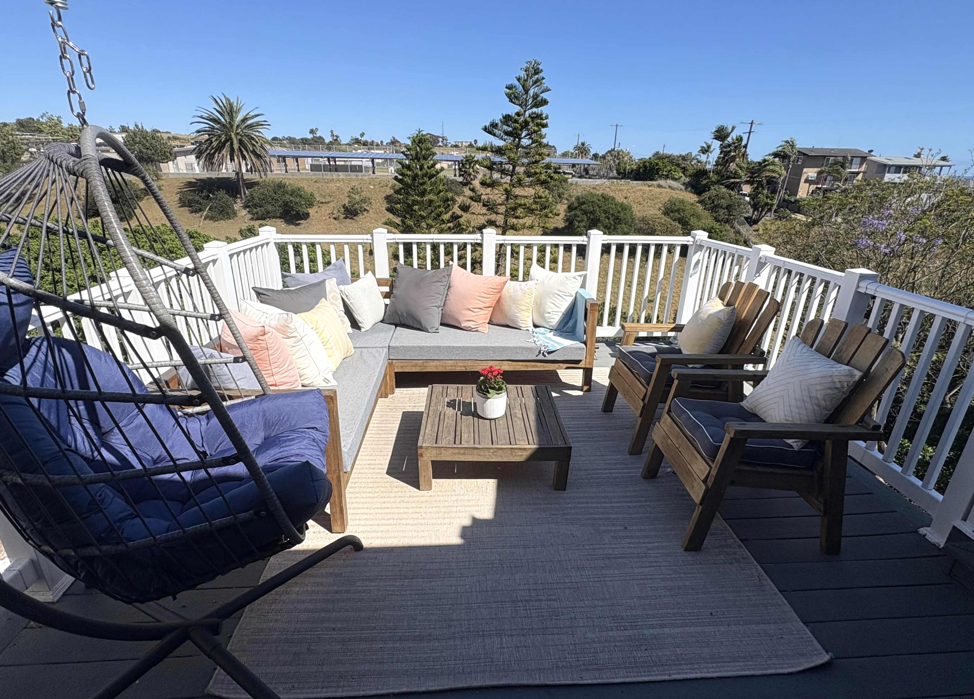 A cozy outdoor seating area features a gray sofa with colorful cushions, a wooden coffee table, and two rustic armchairs, surrounded by a serene landscape and clear blue sky.