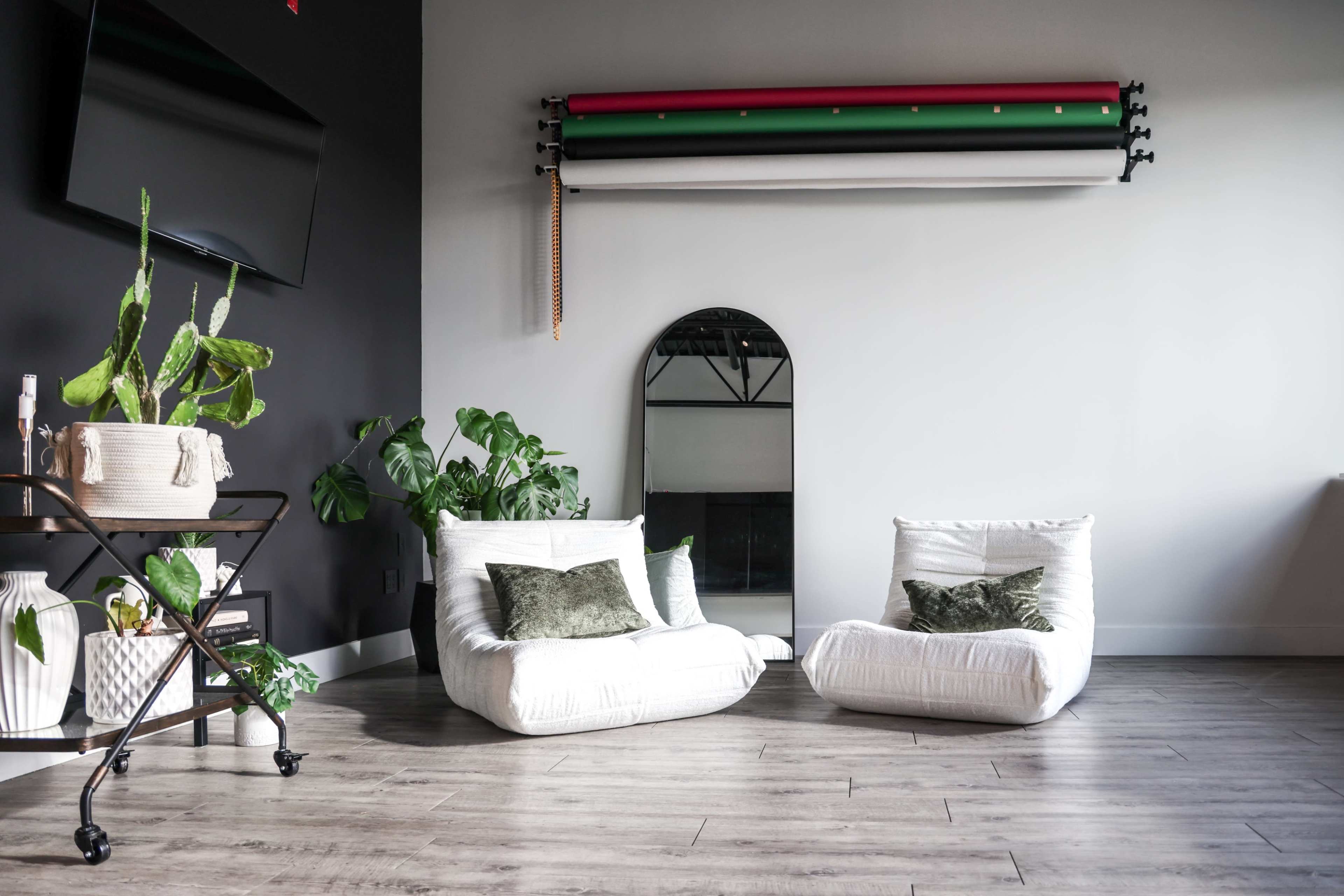 The image shows a modern interior featuring two white lounge chairs, a tall mirror, assorted indoor plants, and a black wall with a TV mounted above.