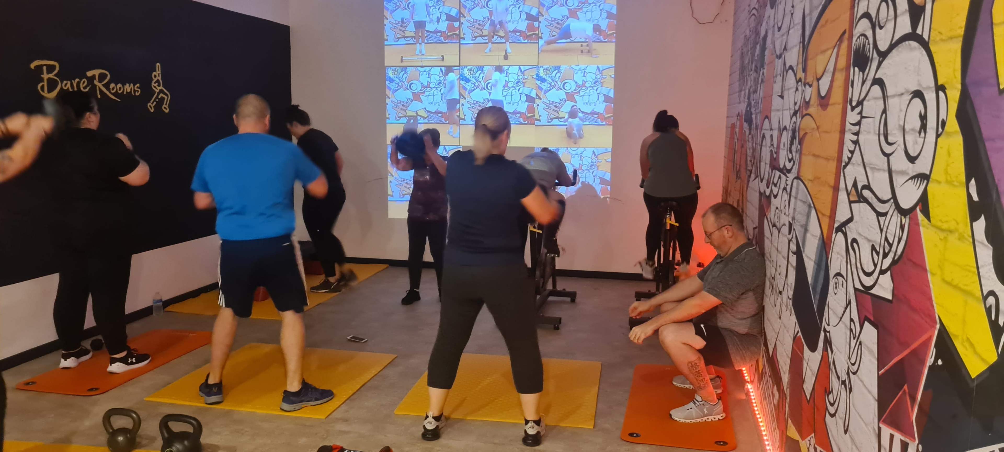 A group of people participates in a fitness class with exercise mats and stationary bikes, while a screen displays animated visuals.