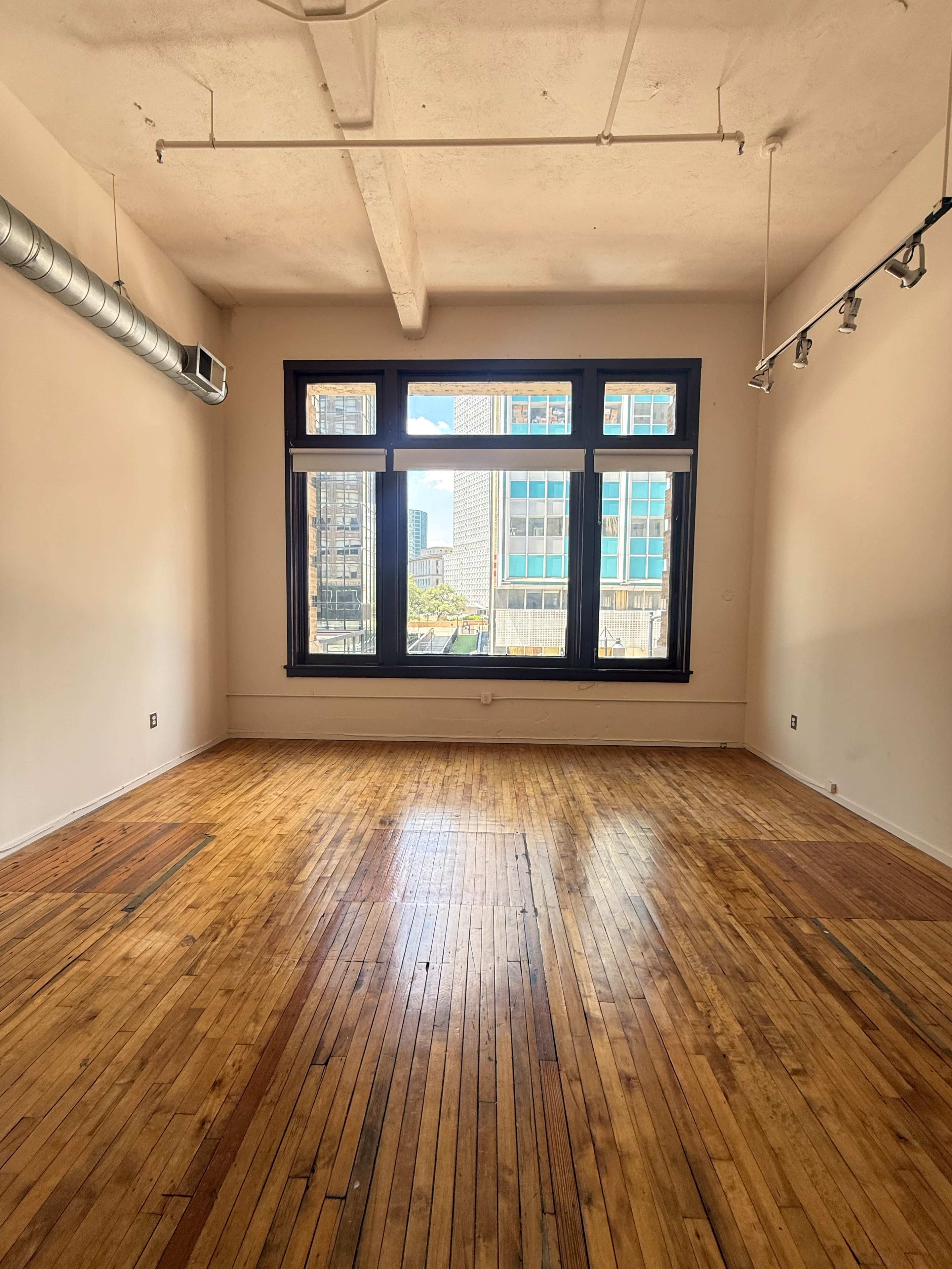 The image shows a spacious, empty room with hardwood floors and large windows overlooking a cityscape.