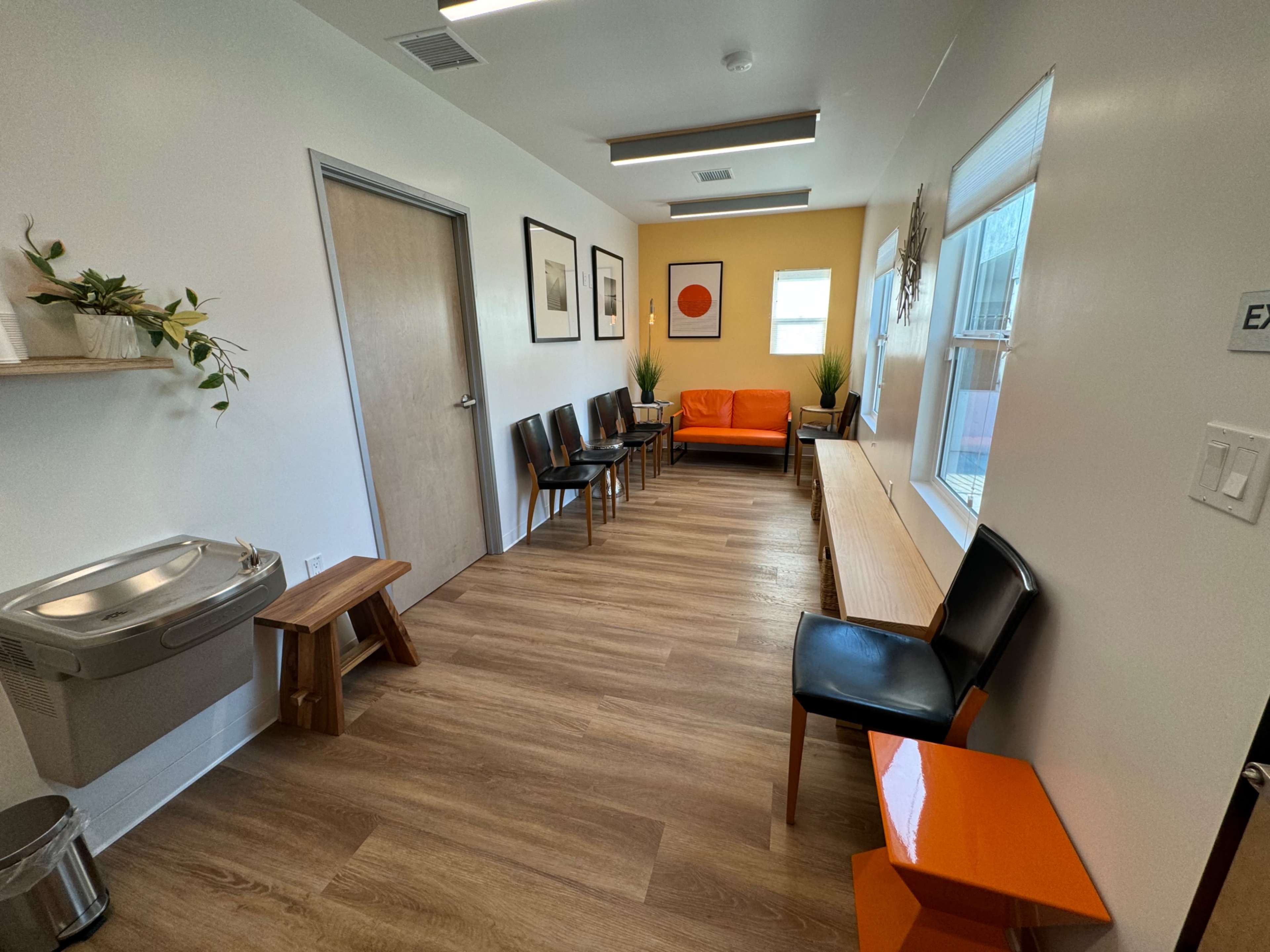A waiting room with several chairs arranged along the walls, a bench, an orange couch, a water fountain, and potted plants.