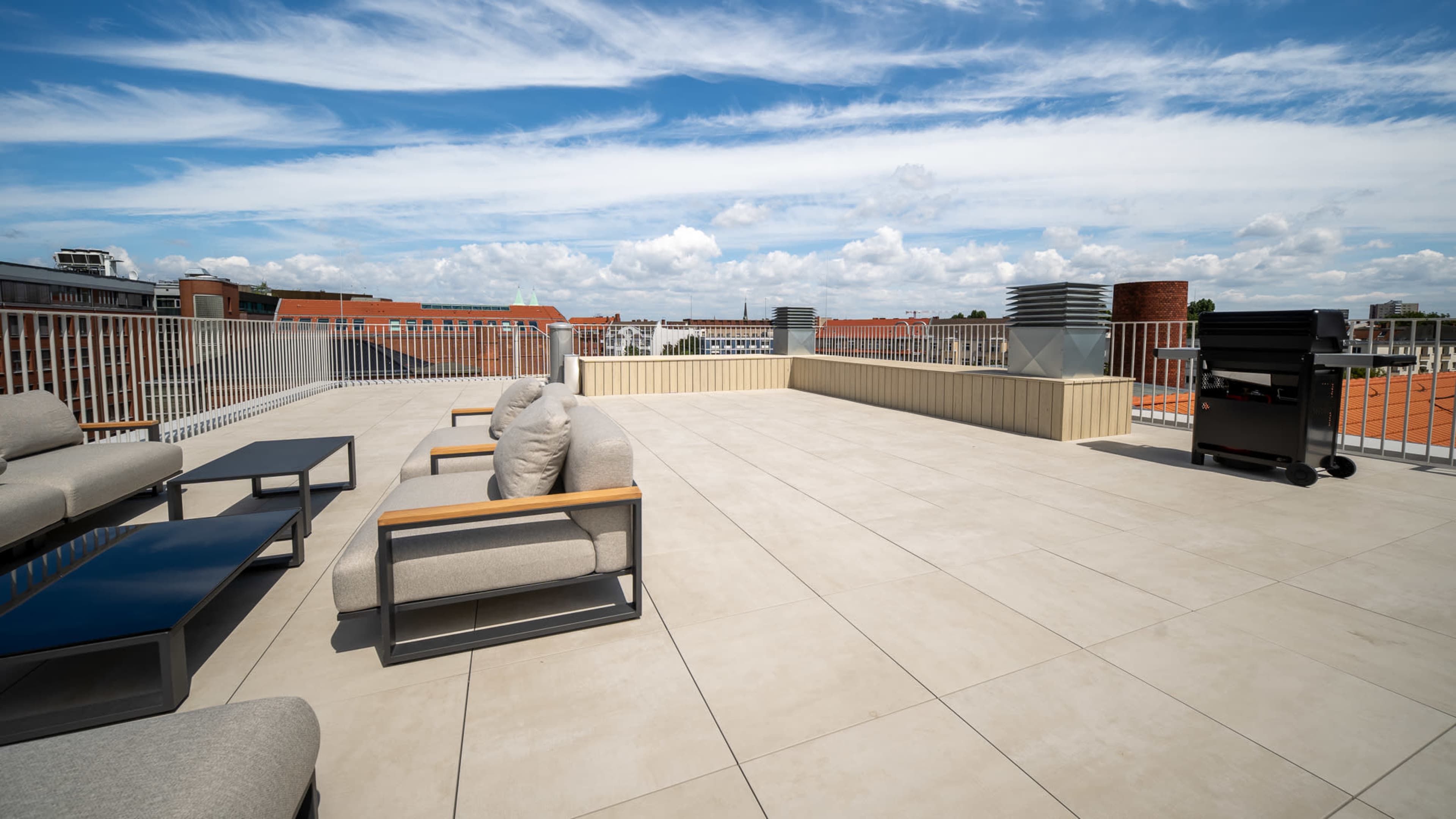 The image shows a spacious rooftop terrace with modern furniture, including couches and tables, and a grill against a backdrop of a clear blue sky.
