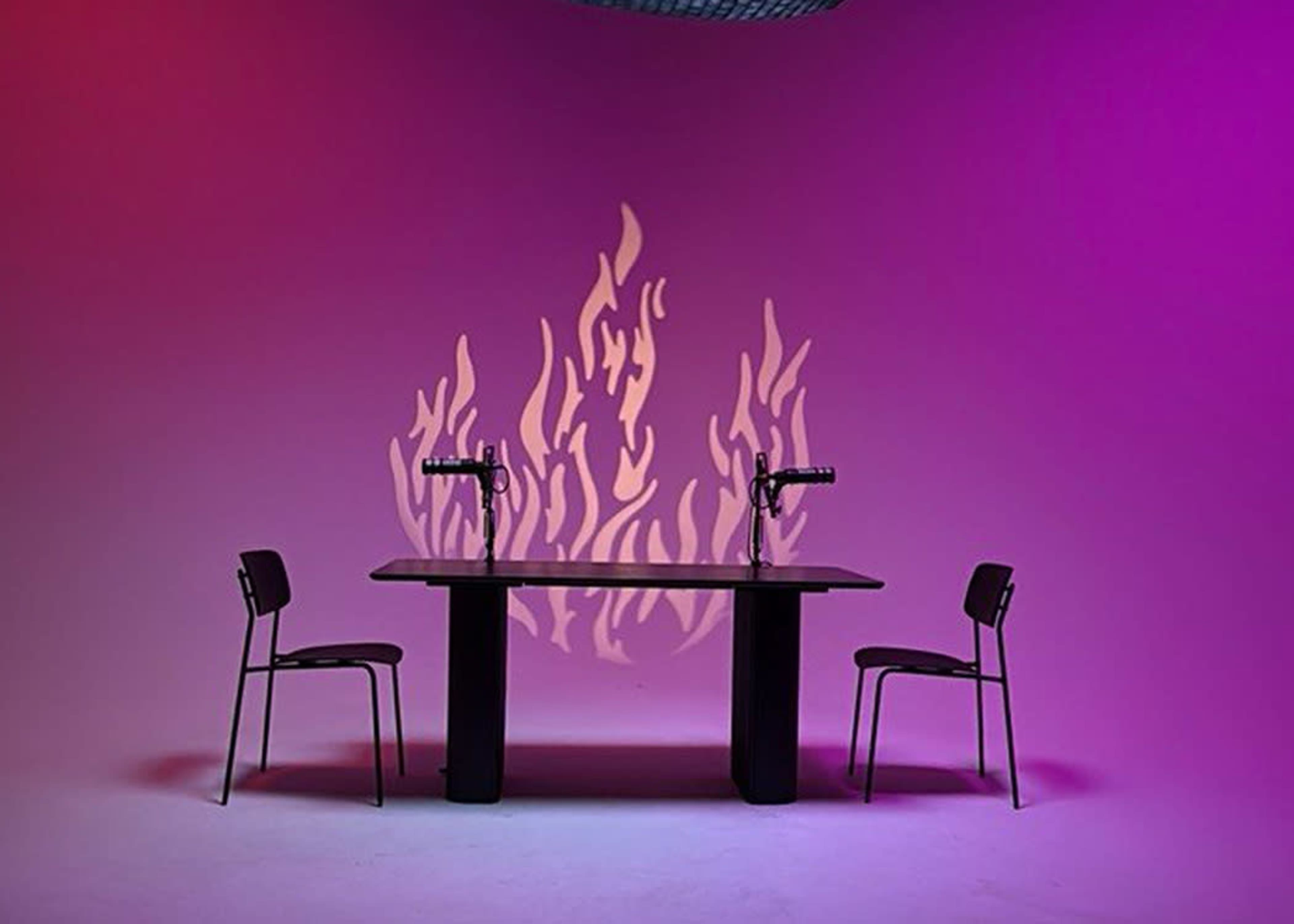 A table with two chairs is set against a purple backdrop, featuring a shadow of flames projected on the wall.