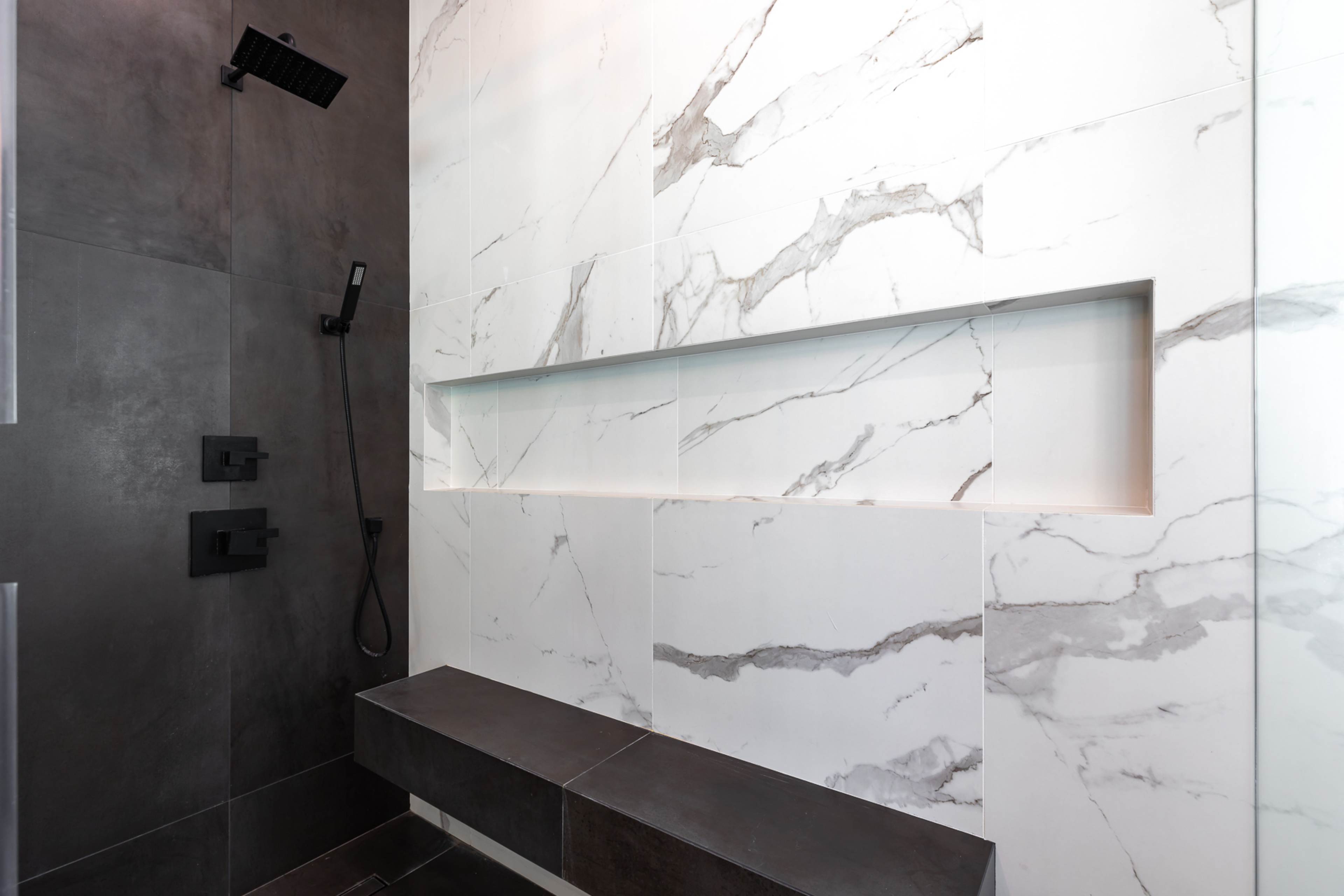 A modern shower with a dark tile floor, marble walls, and a built-in shelf.