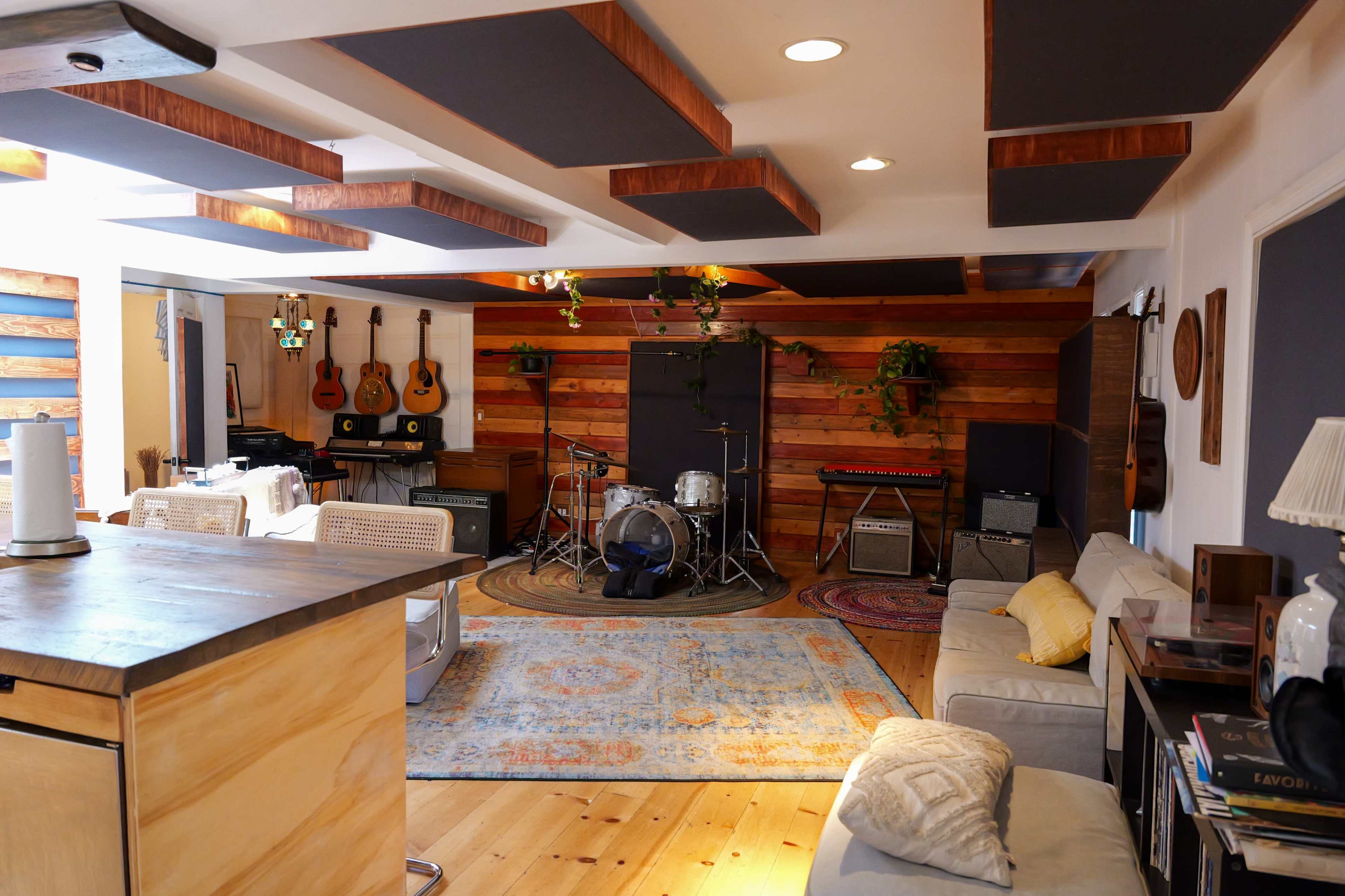 A music studio features a drum set and various musical instruments arranged against wooden walls, with comfortable seating and a patterned rug on the floor.