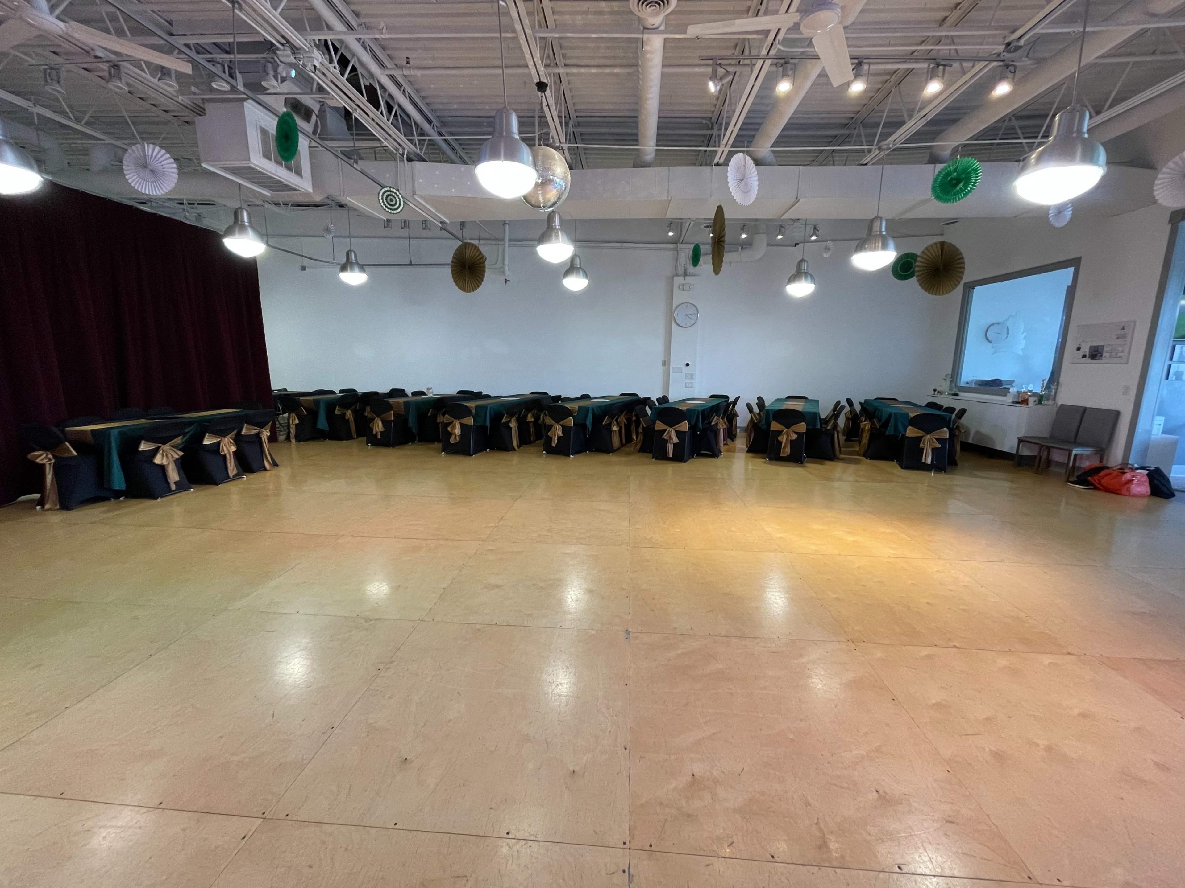 A spacious event room features several tables covered in dark cloths with gold sashes, arranged in a U-shape, under overhead lights.