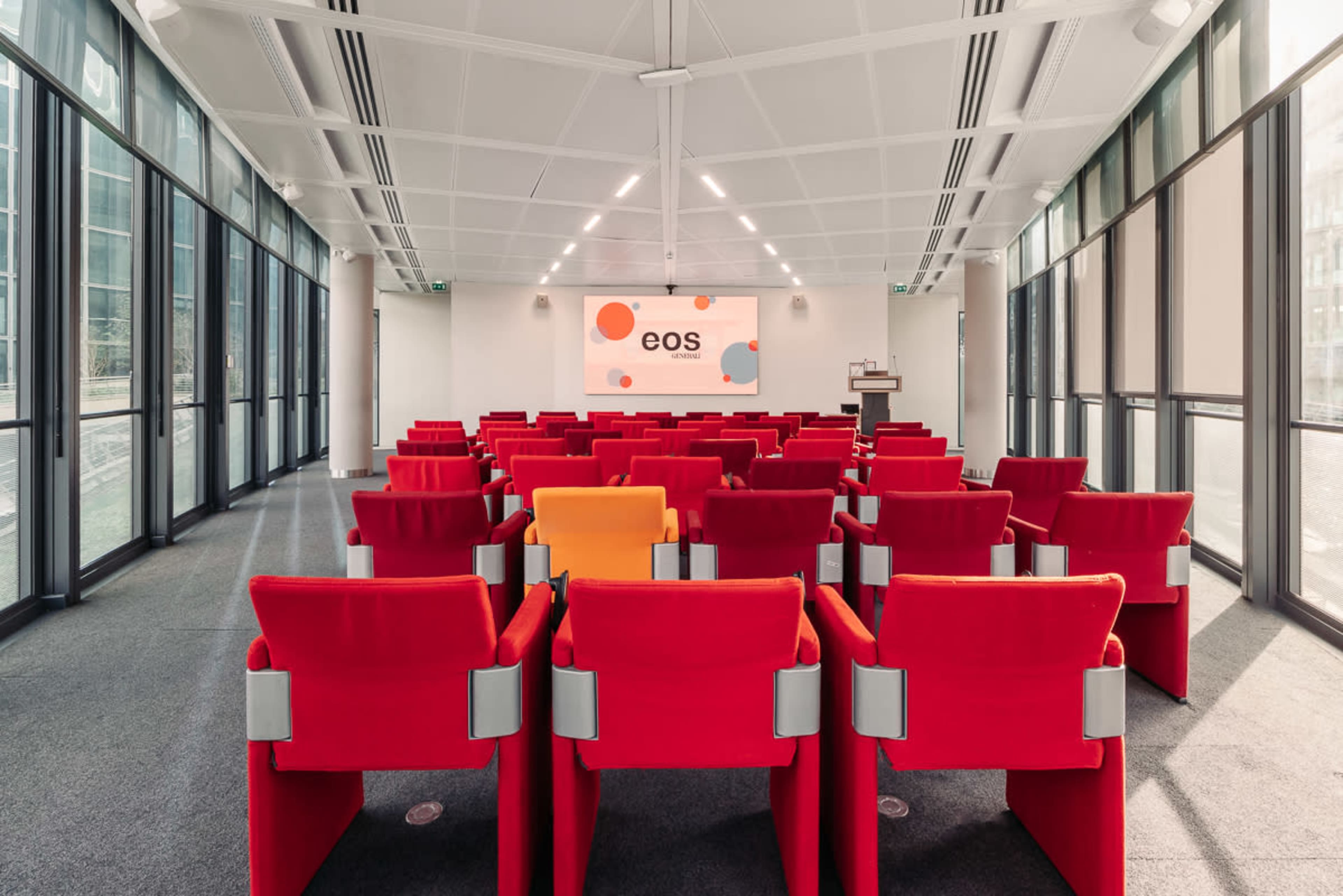 The image shows a modern conference room with rows of red chairs facing a large screen that displays the word "eos."