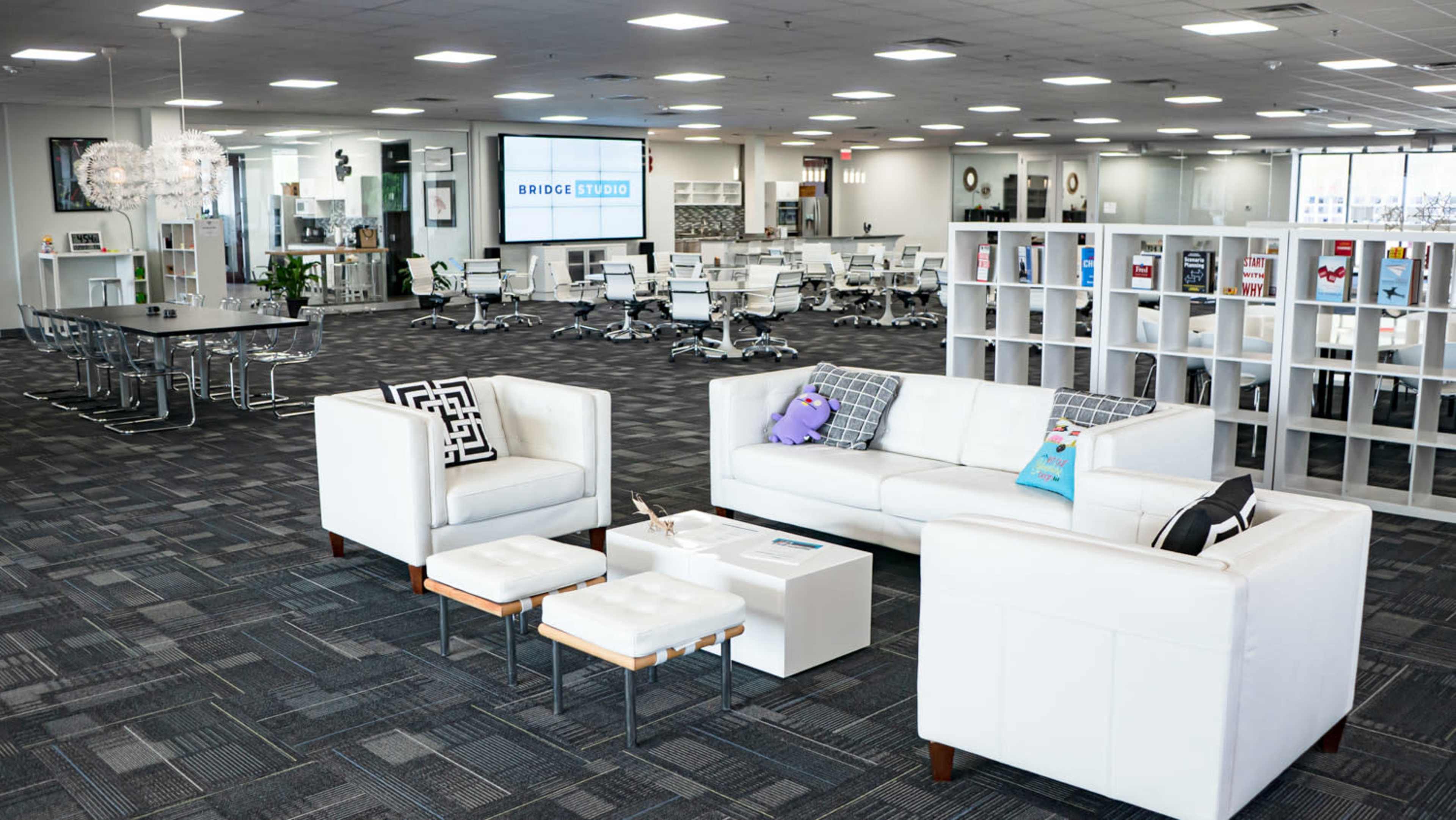 A modern office space with a seating area featuring a white sofa and small tables, surrounded by workstations and large screens displaying a logo.