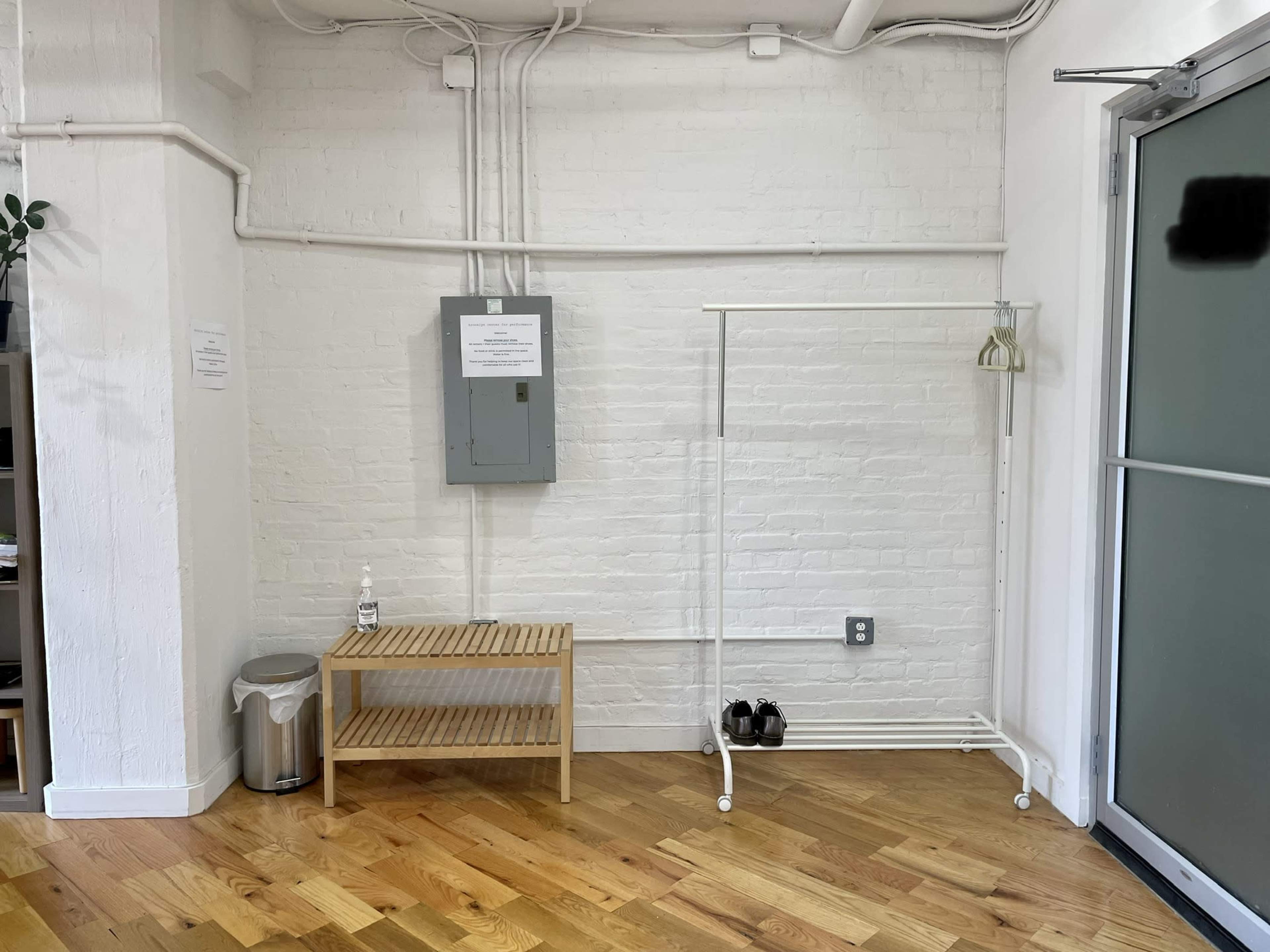 A small, minimalist room with a wooden bench, a clothes rack, a trash can, and electrical panel mounted on a white brick wall.