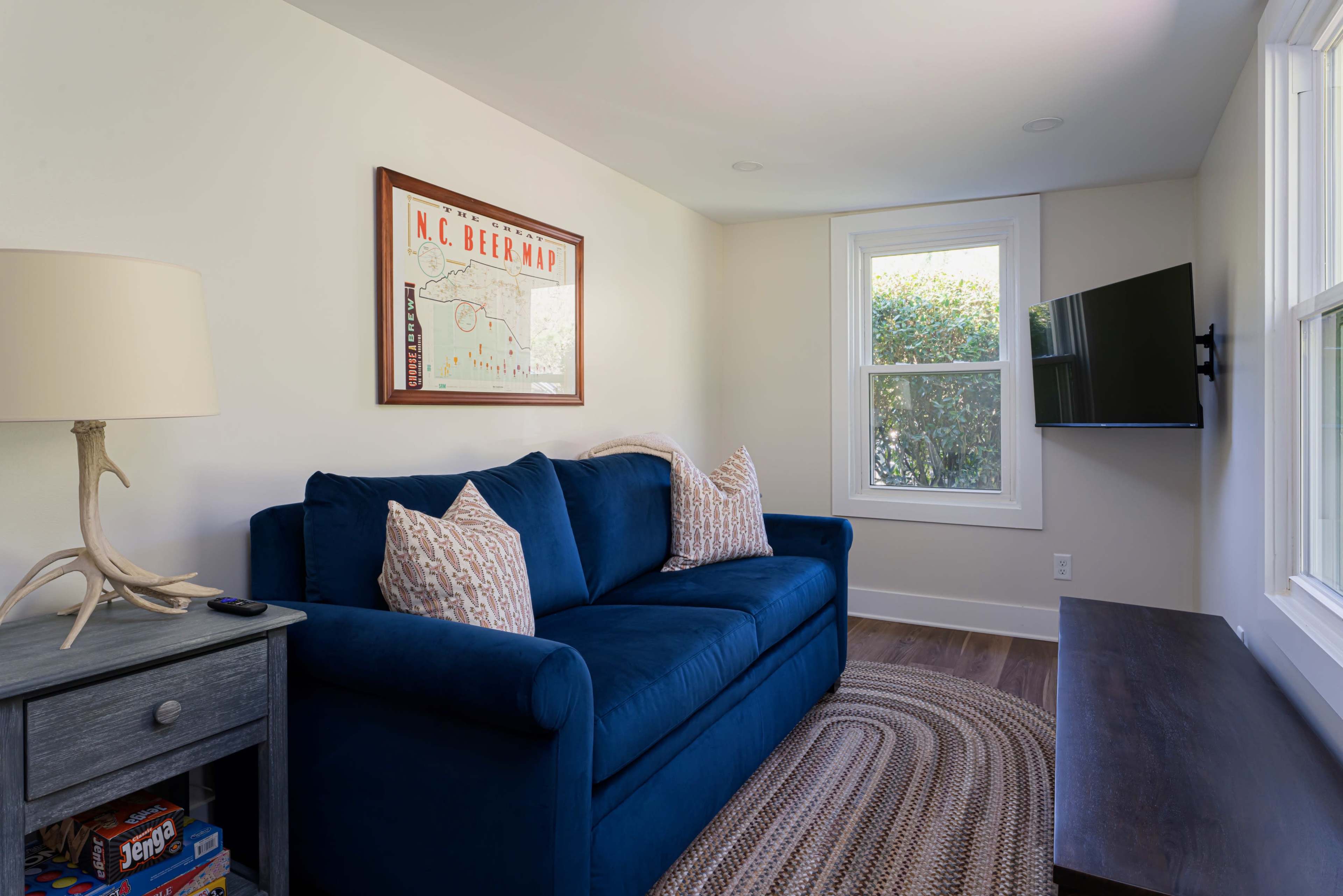 The image shows a cozy living area featuring a blue couch with patterned pillows, a wall-mounted TV, a wooden side table with a lamp, and framed artwork on the wall.