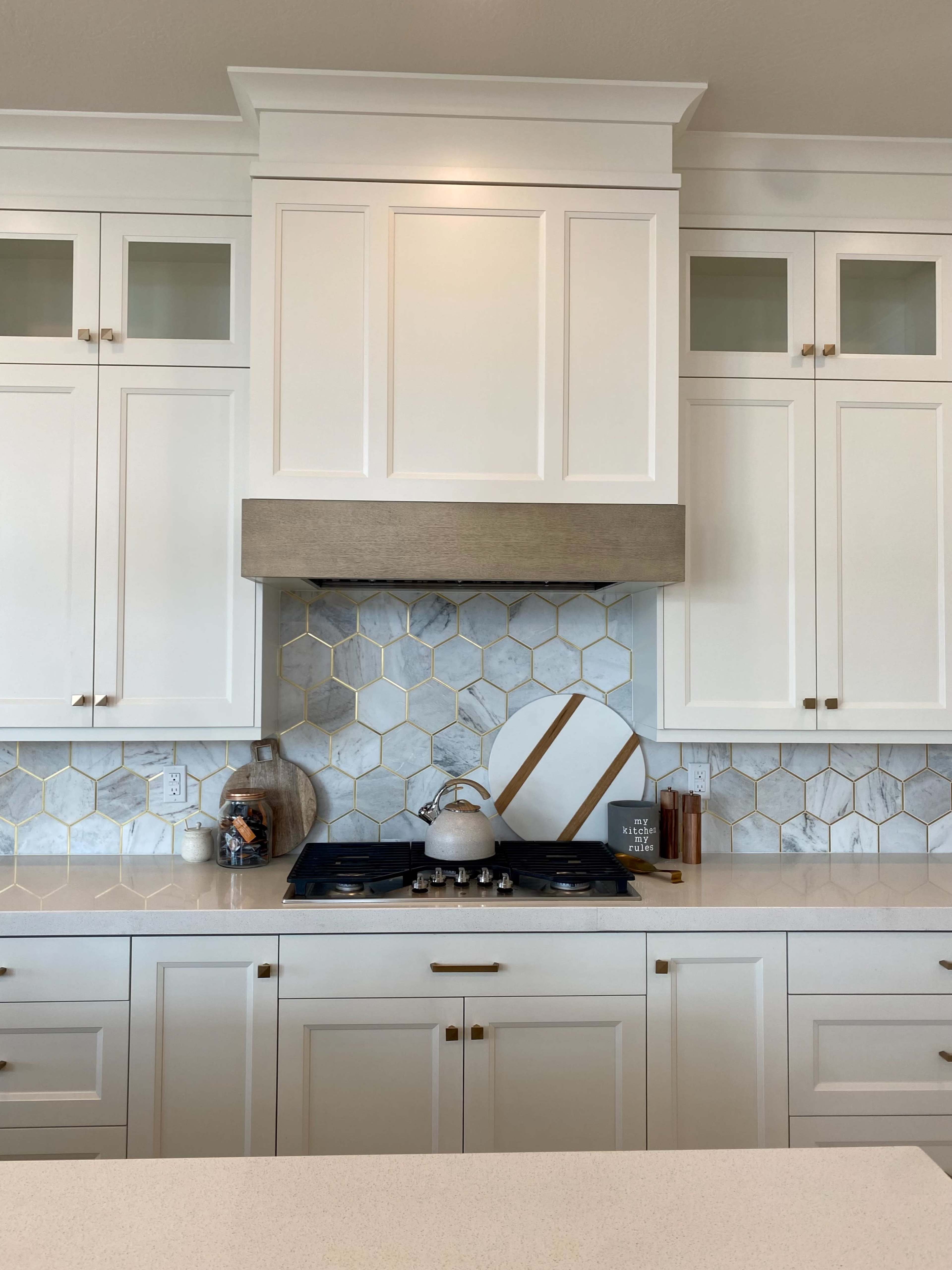 The image shows a modern kitchen with white cabinetry, a marble backsplash featuring hexagonal tiles, and a gas cooktop beneath a vent hood.
