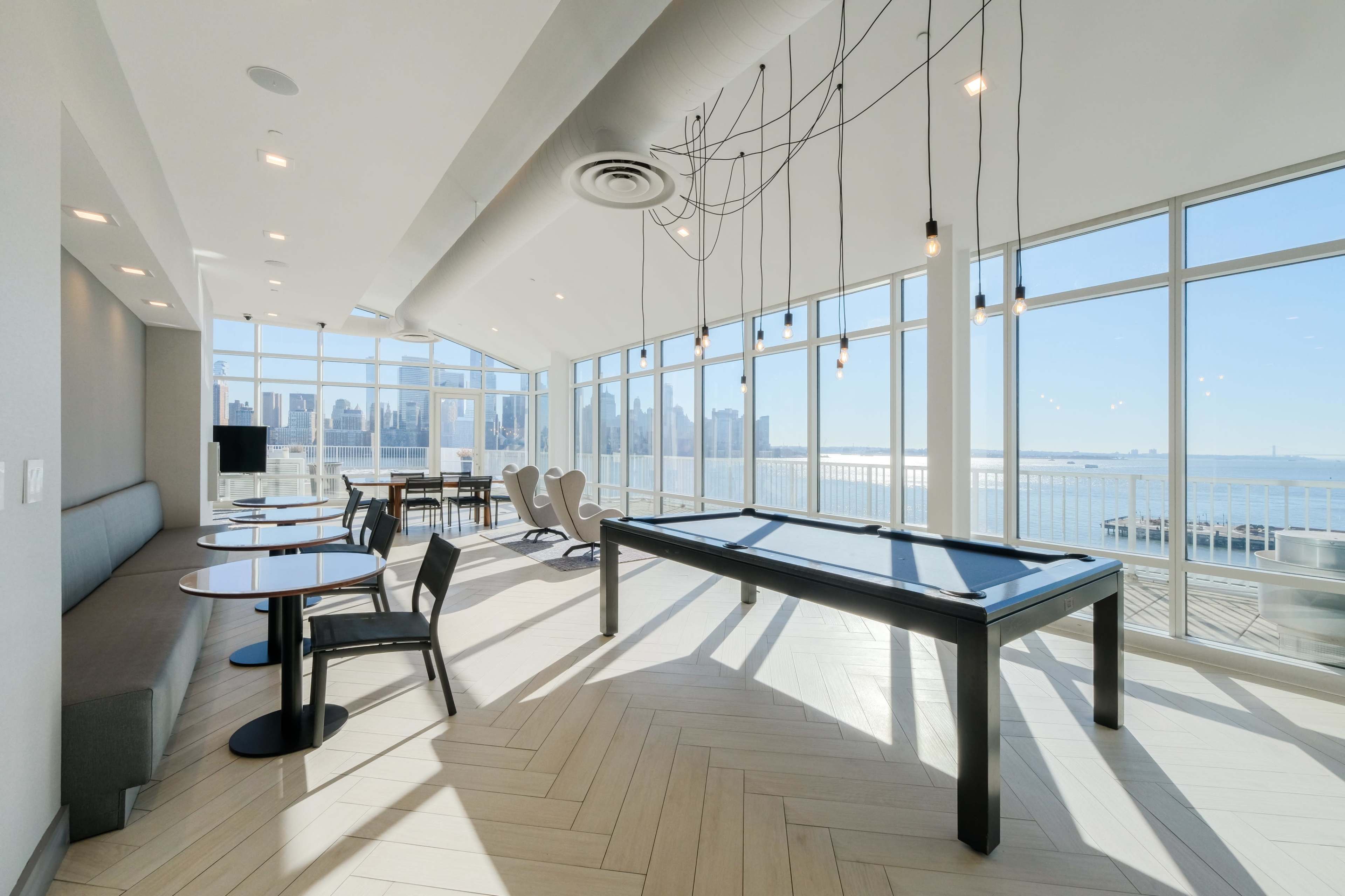 The image shows a modern room with large windows overlooking a city skyline, featuring a billiard table and several seating areas.