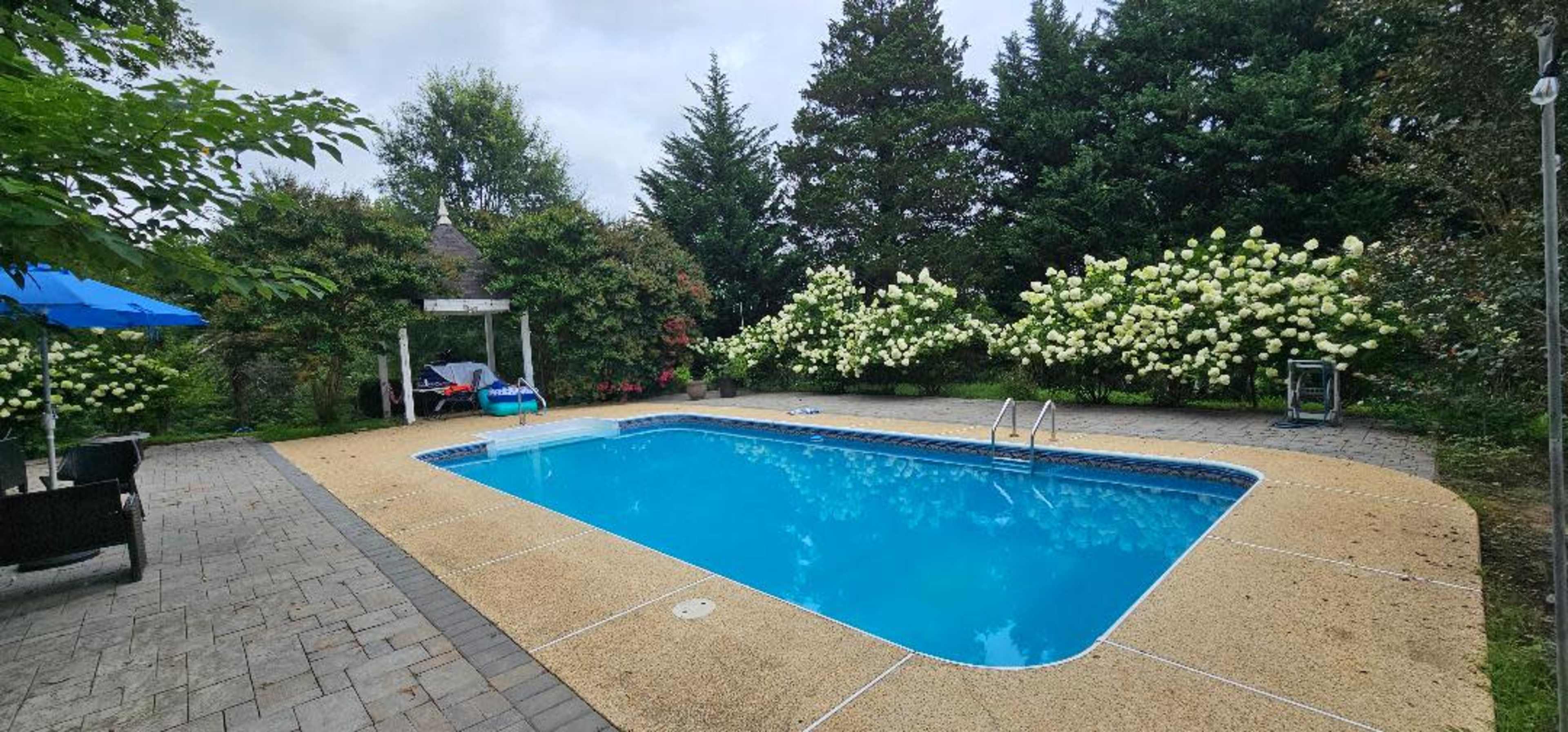 A rectangular swimming pool surrounded by flower-filled hedges and a stone patio, with garden furniture and an umbrella nearby.