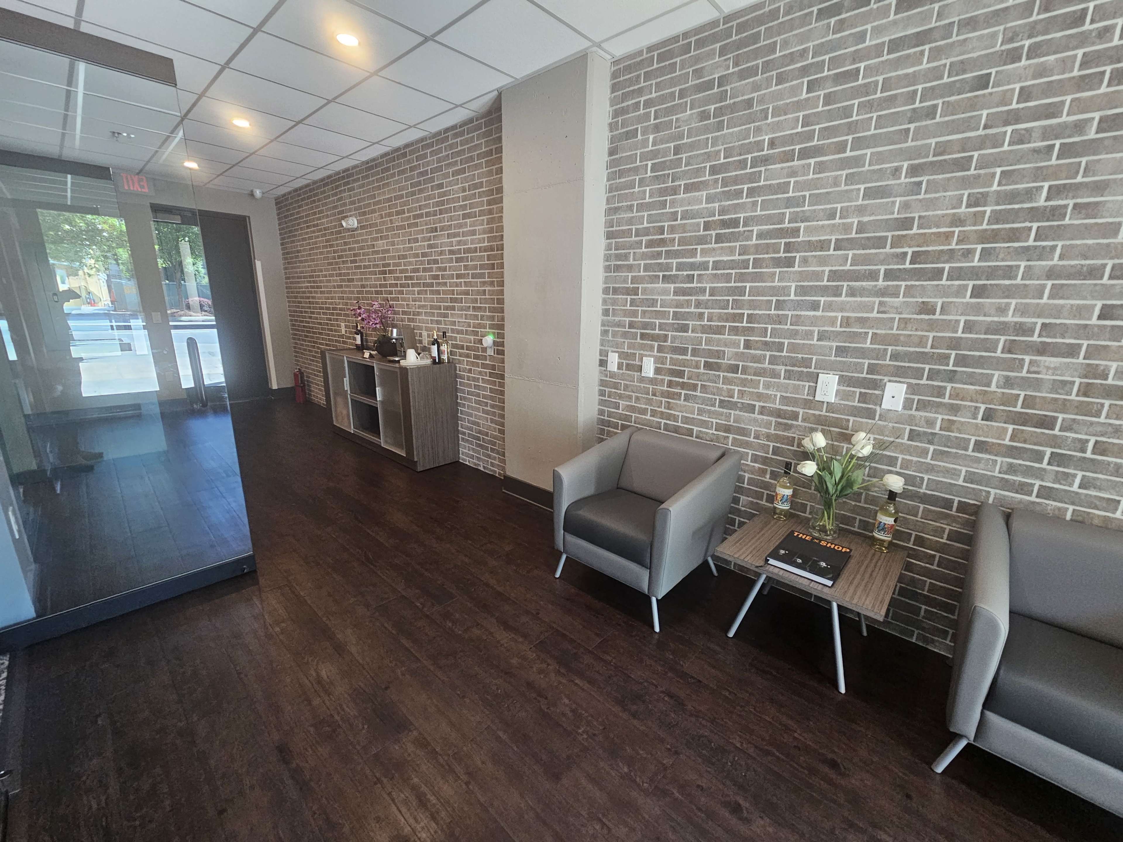 The image shows a modern reception area featuring a brick wall, two gray armchairs, a wooden table with decorative items, and a glass entrance with natural light coming in.