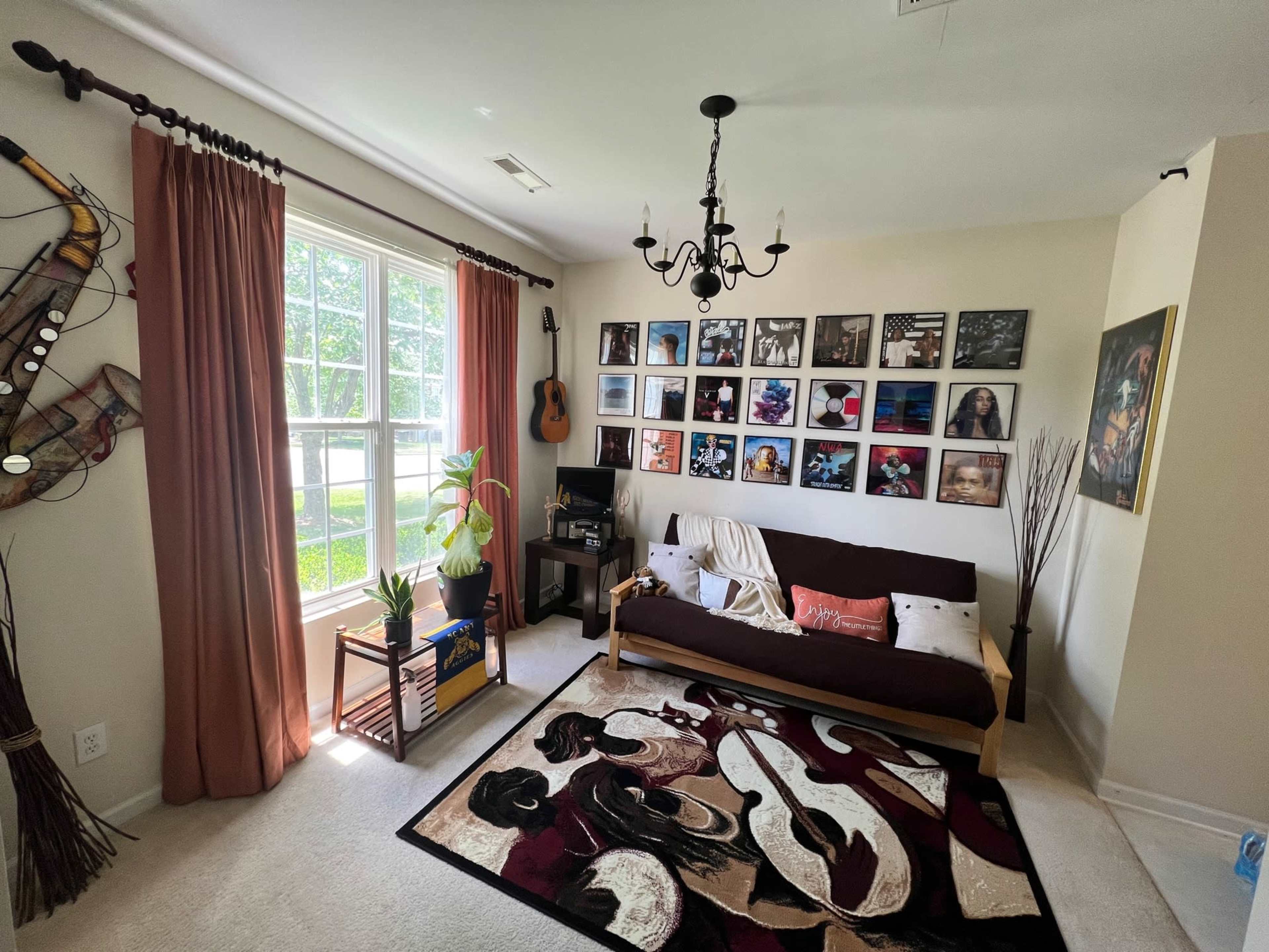 A cozy living room with a sofa bed, a colorful area rug, a wall of framed photographs, and large windows with curtains.