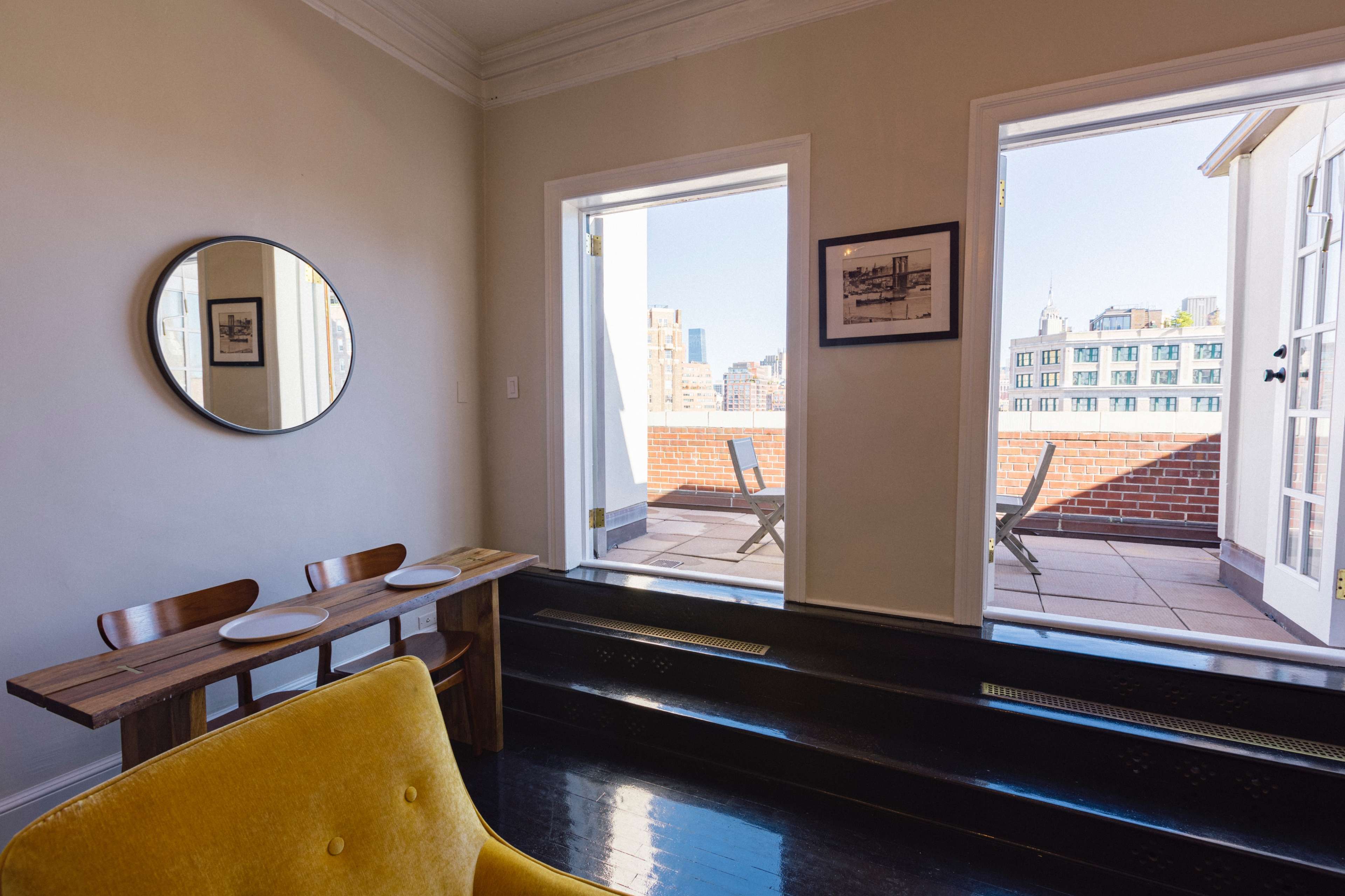 A bright room features a yellow chair, a wooden table set for two, and two open doors leading to a rooftop terrace.