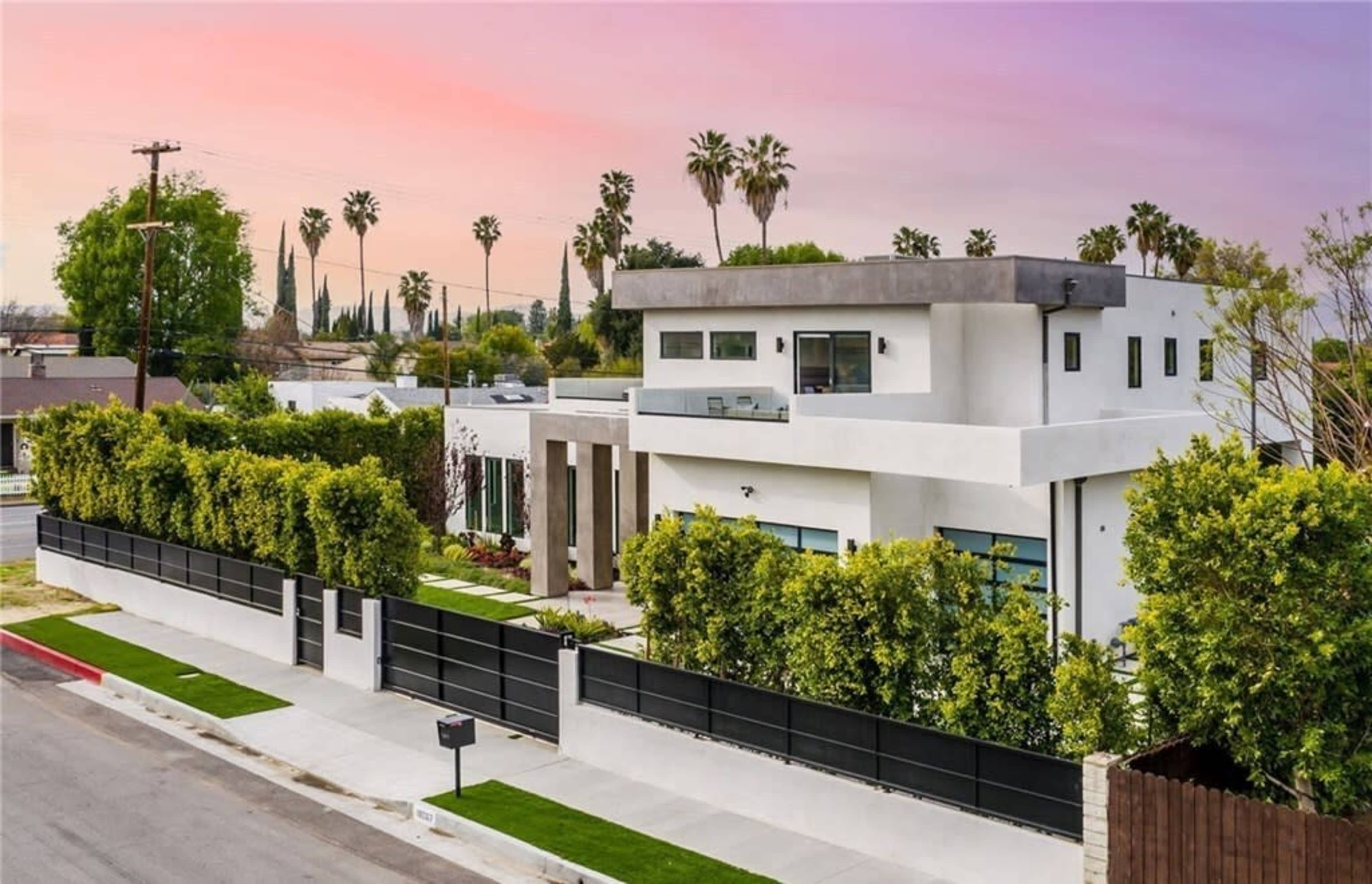 A modern two-story house with a flat roof and large windows is surrounded by greenery and features a black fence along the front.