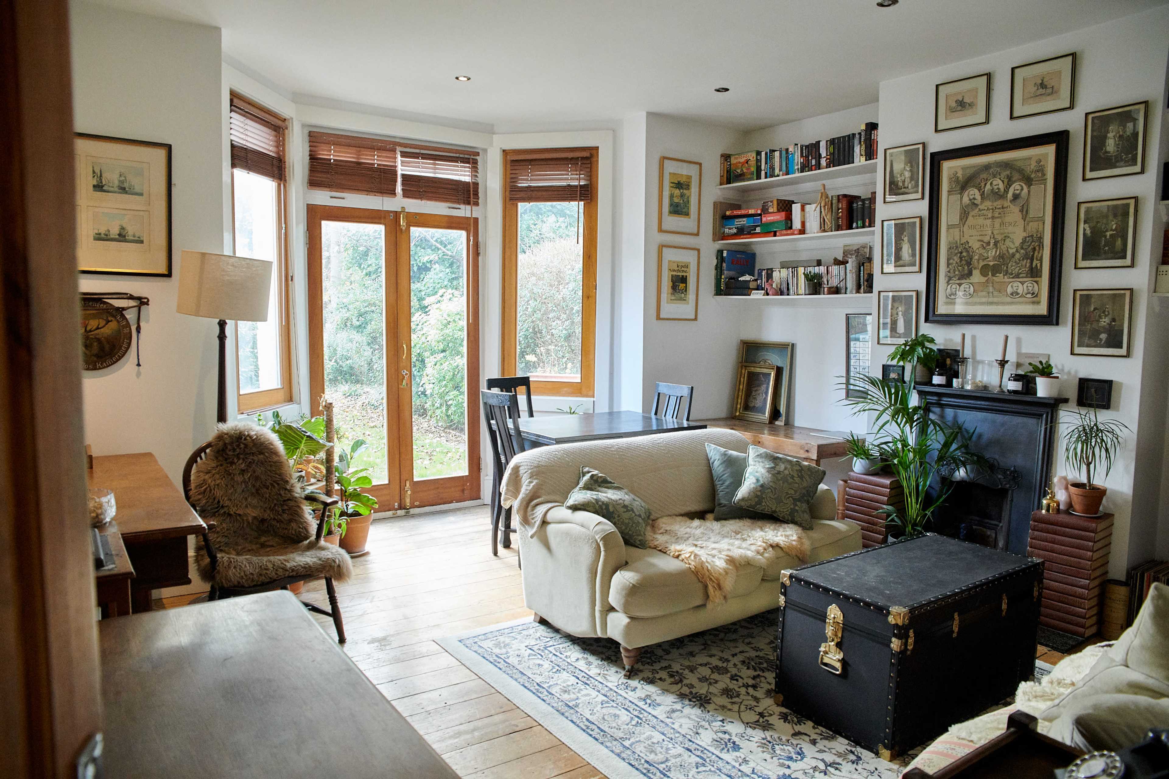 A cozy living room features a mix of bookshelves, a fireplace, and large windows overlooking a garden.