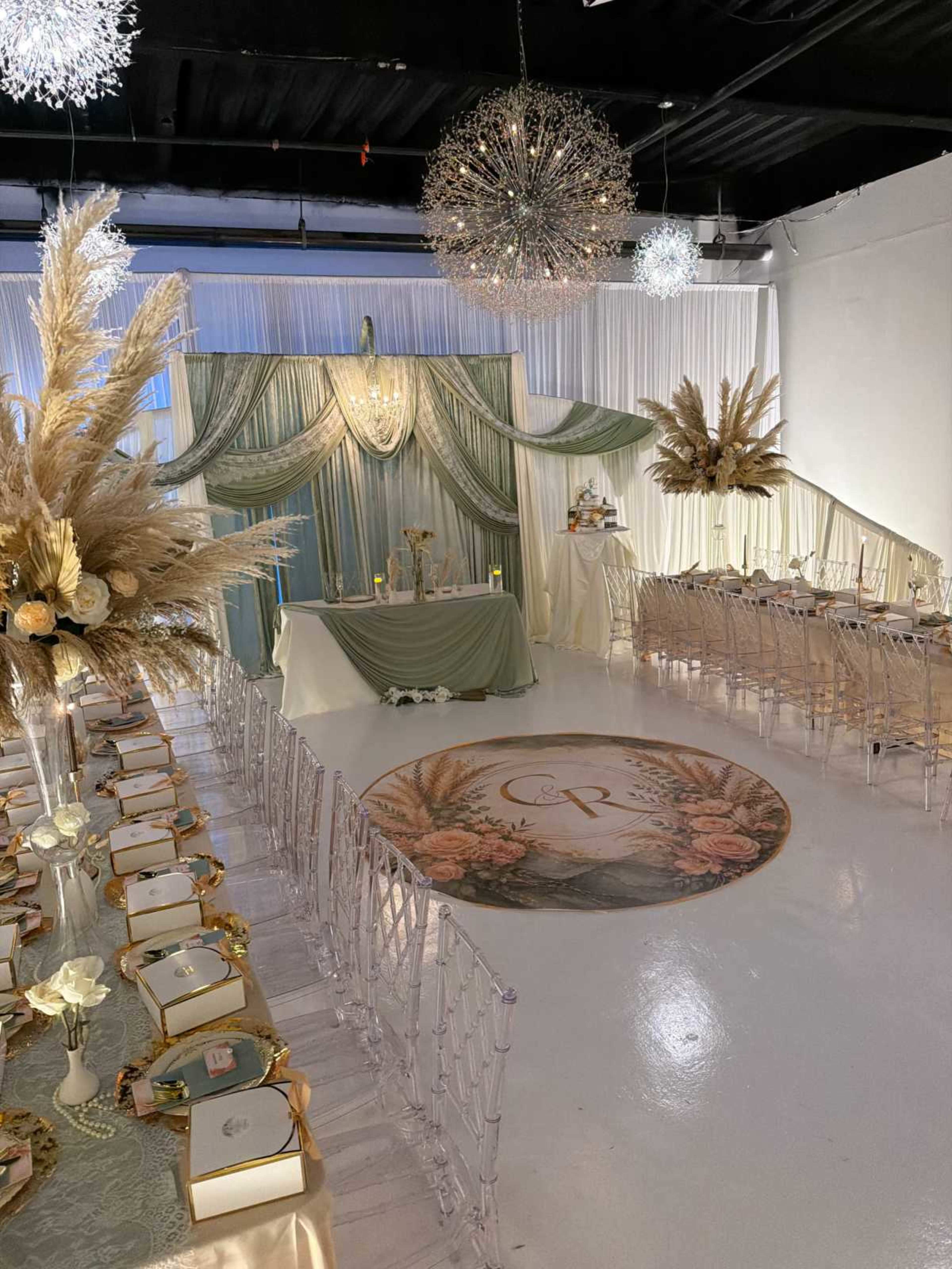 The image shows a elegantly arranged event space with a central backdrop, decorative table settings, and a circular logo on the floor.