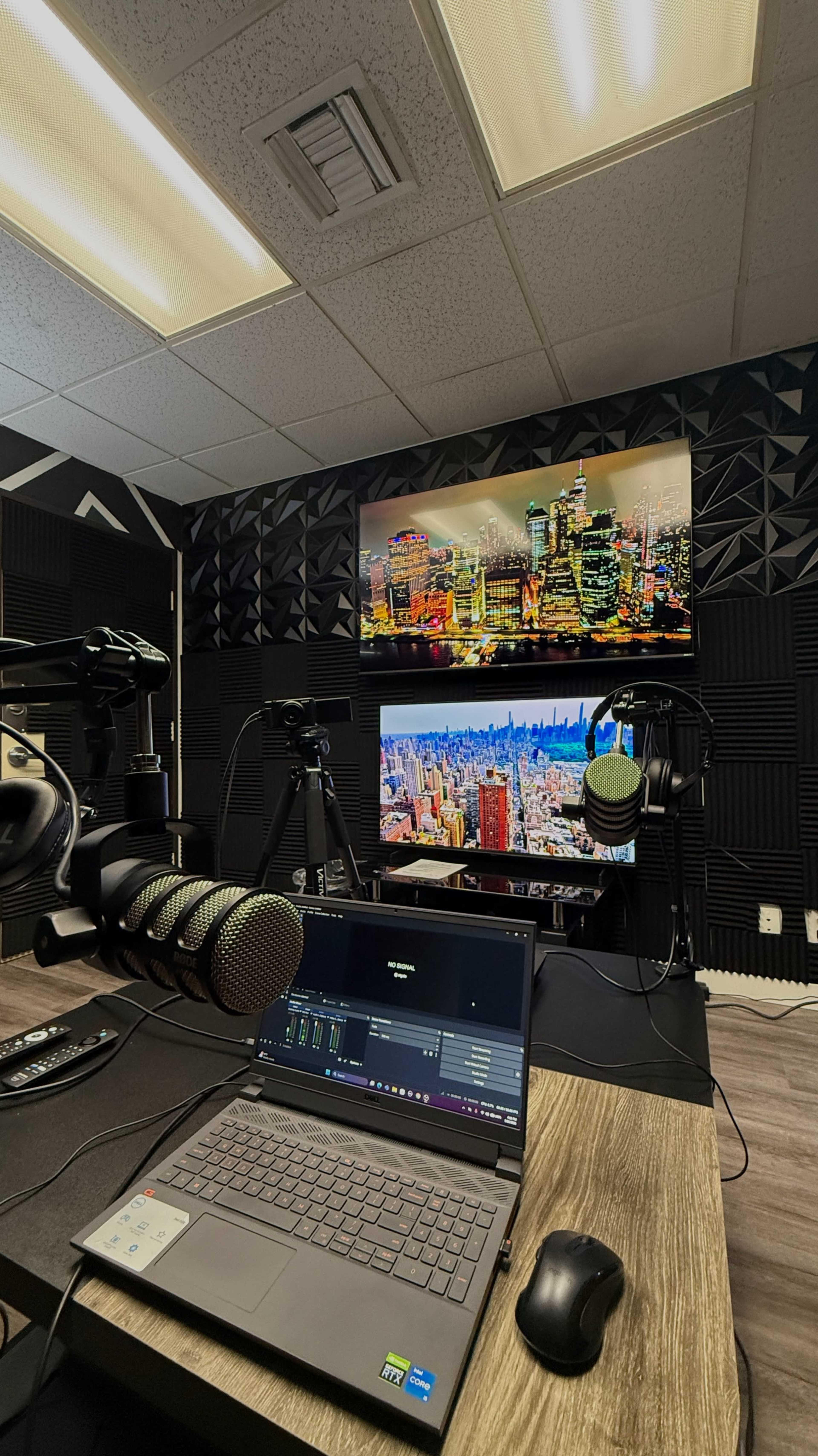 The image shows a recording studio with two large screens displaying cityscapes, a laptop with audio software, and microphones positioned on a wooden desk.