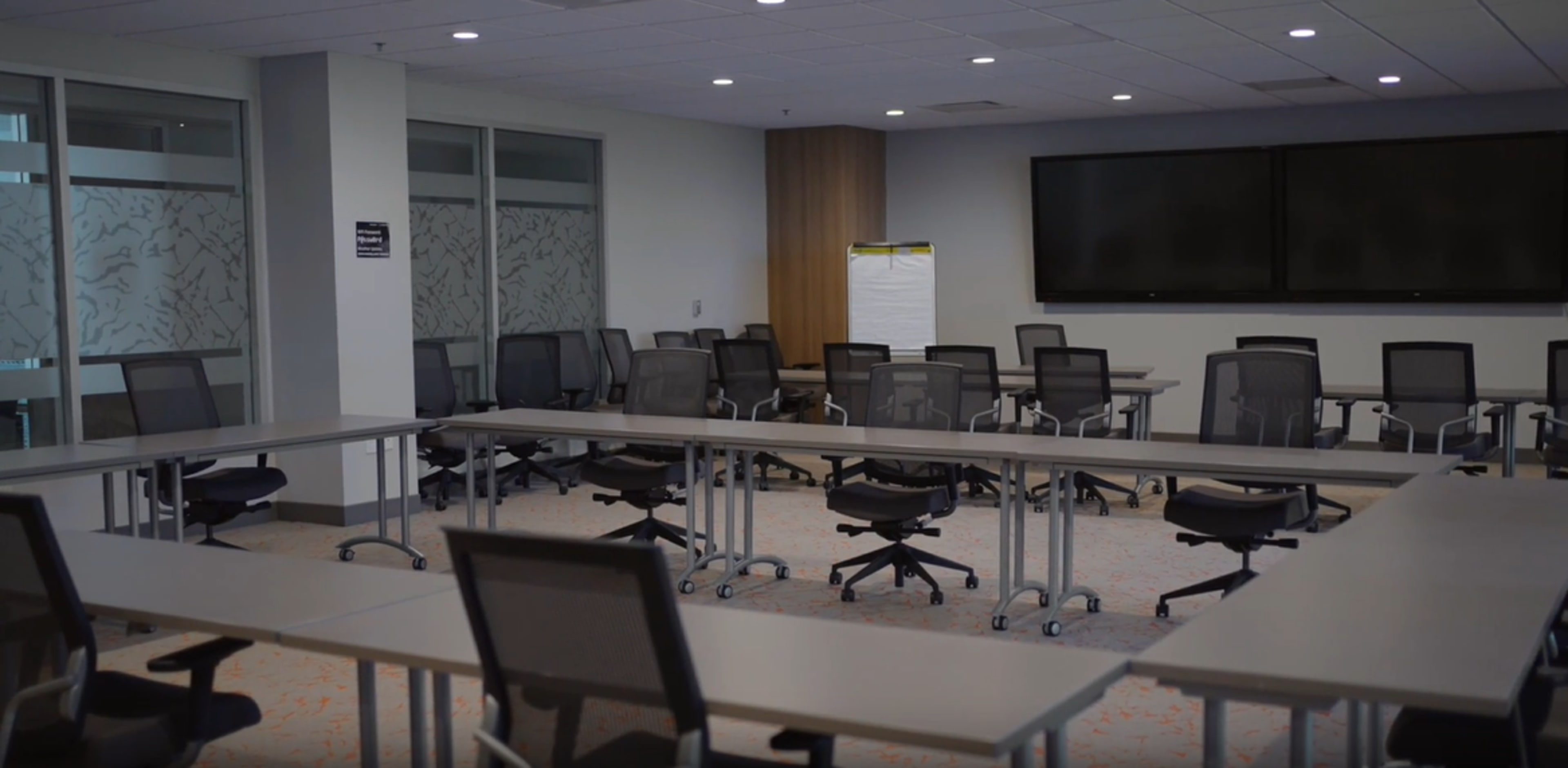 A modern conference room features multiple long tables arranged with ergonomic chairs and a large screen on the wall.