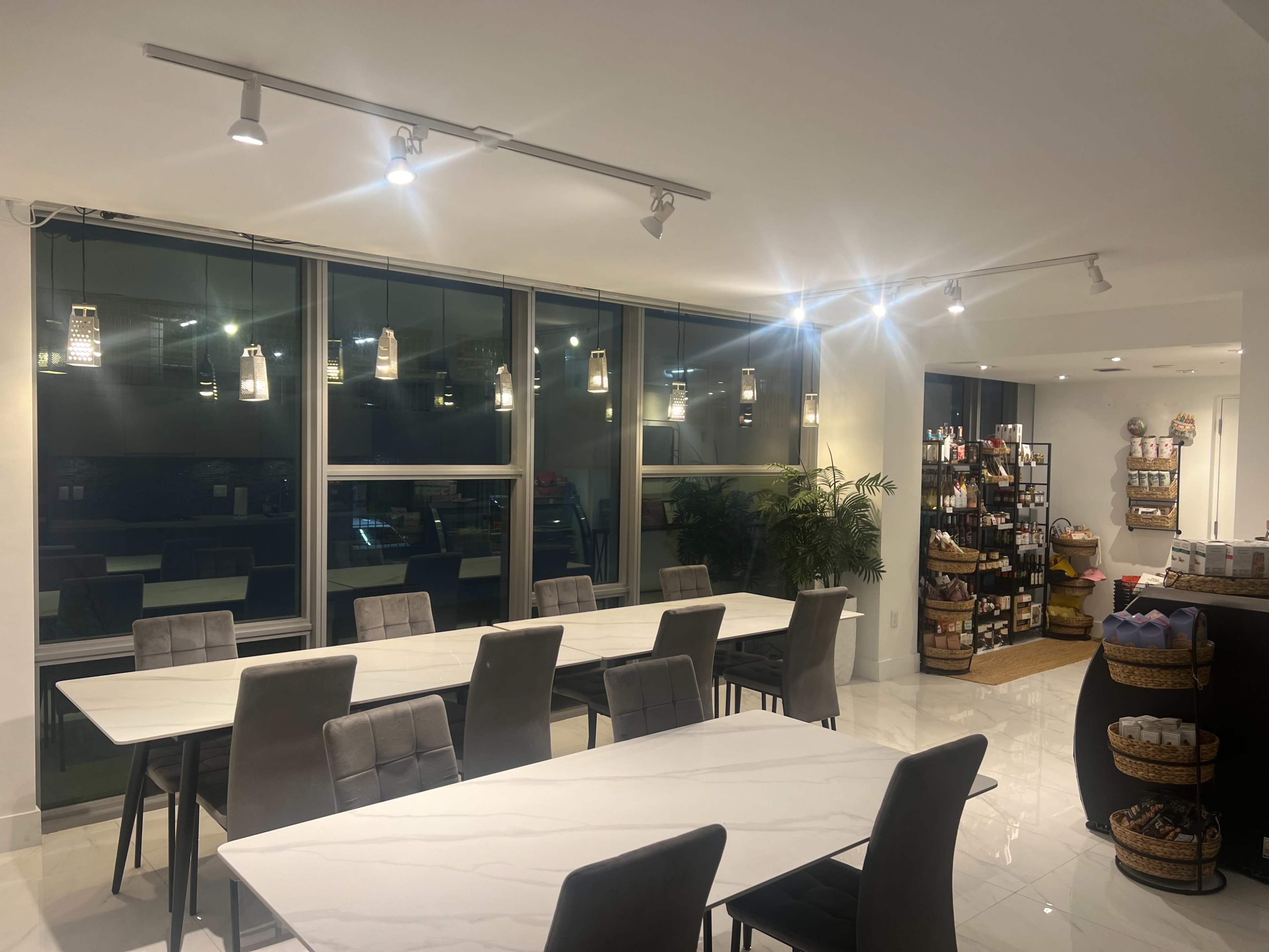 A modern dining area with long tables, gray chairs, and bright overhead lights, featuring a shelf of various food items and products in the background.