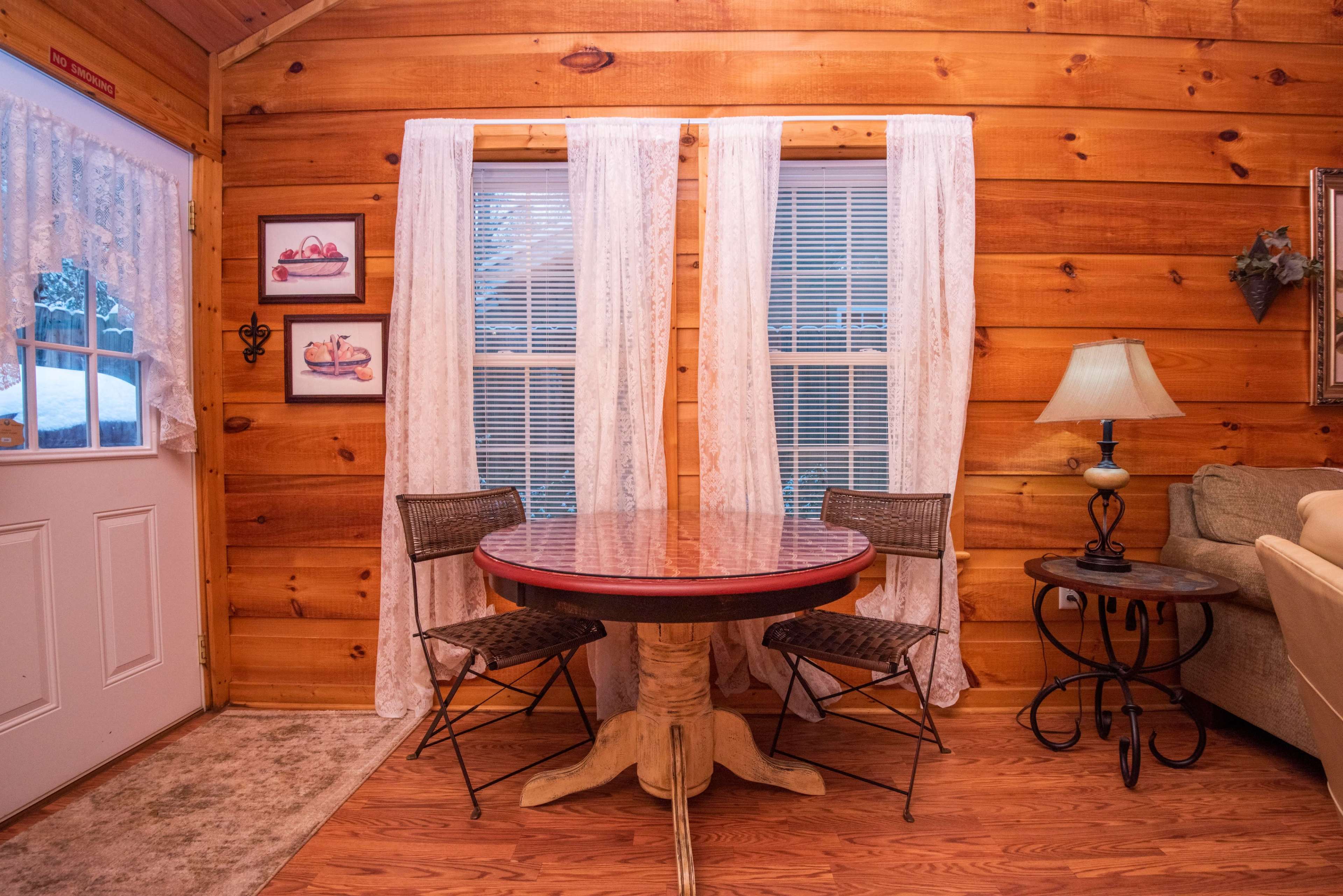 A small wooden table with two chairs is positioned near a door and windows, surrounded by wooden walls in a cozy room.
