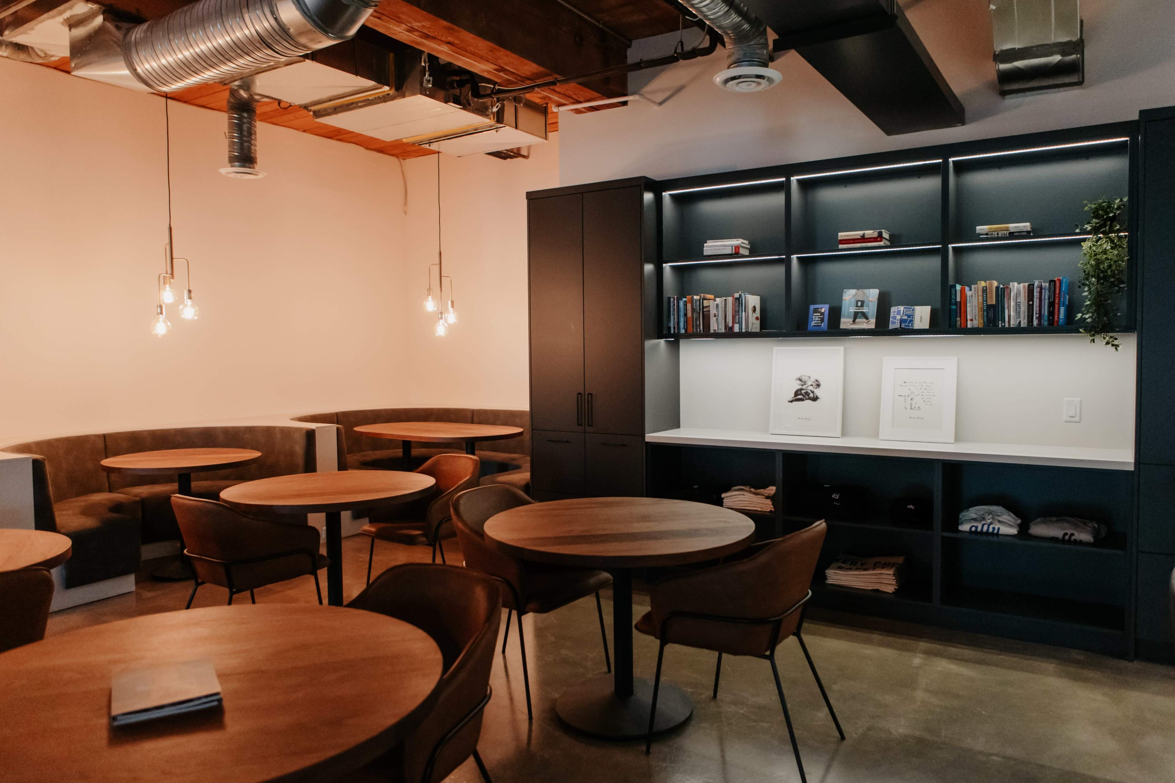 The image features a modern interior space with round wooden tables, upholstered seating along the walls, and a dark shelving unit displaying books and decor.