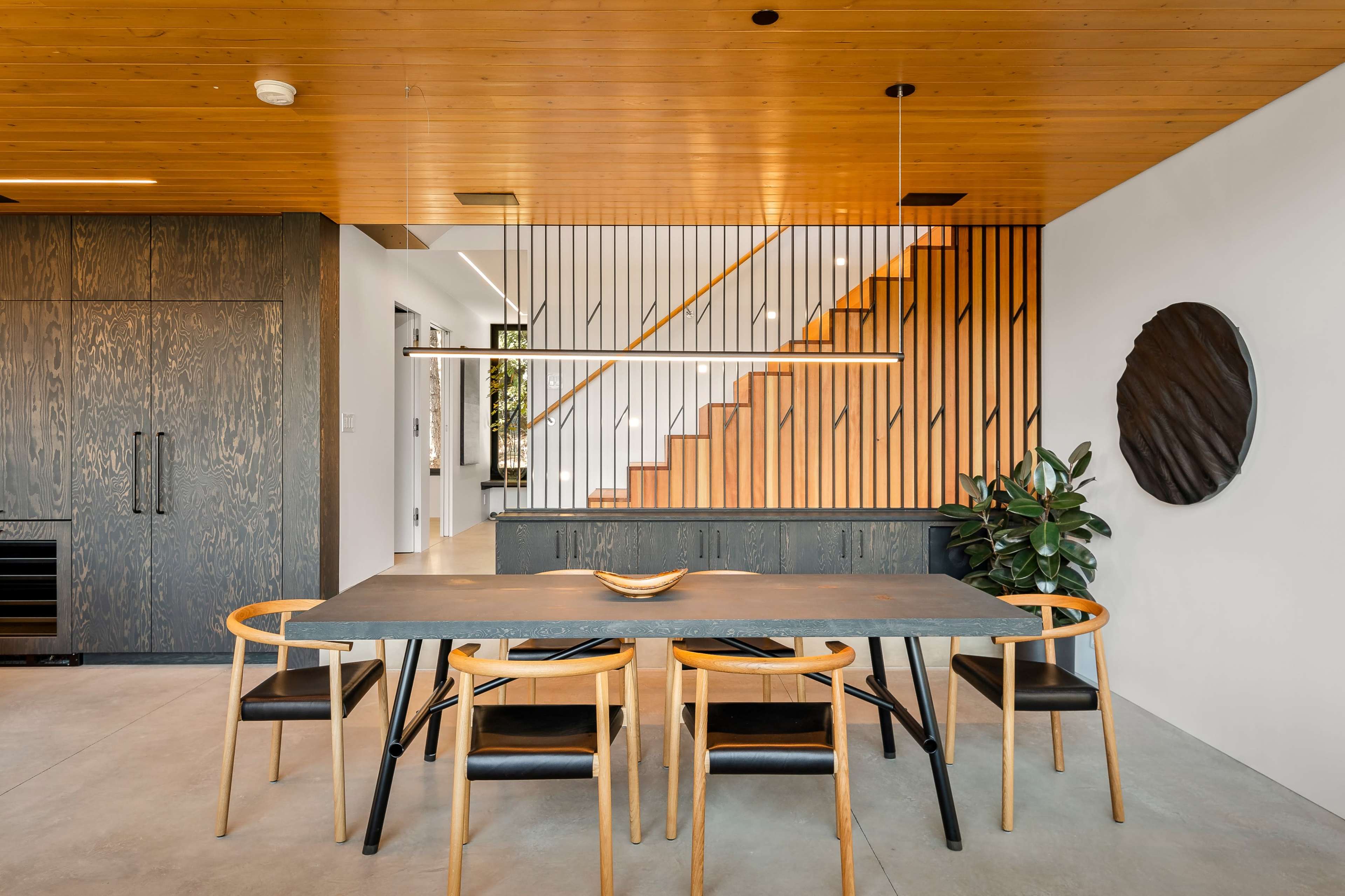 The image displays a modern dining area featuring a rectangular table surrounded by black and wood chairs, with a staircase and a textured wall in the background.