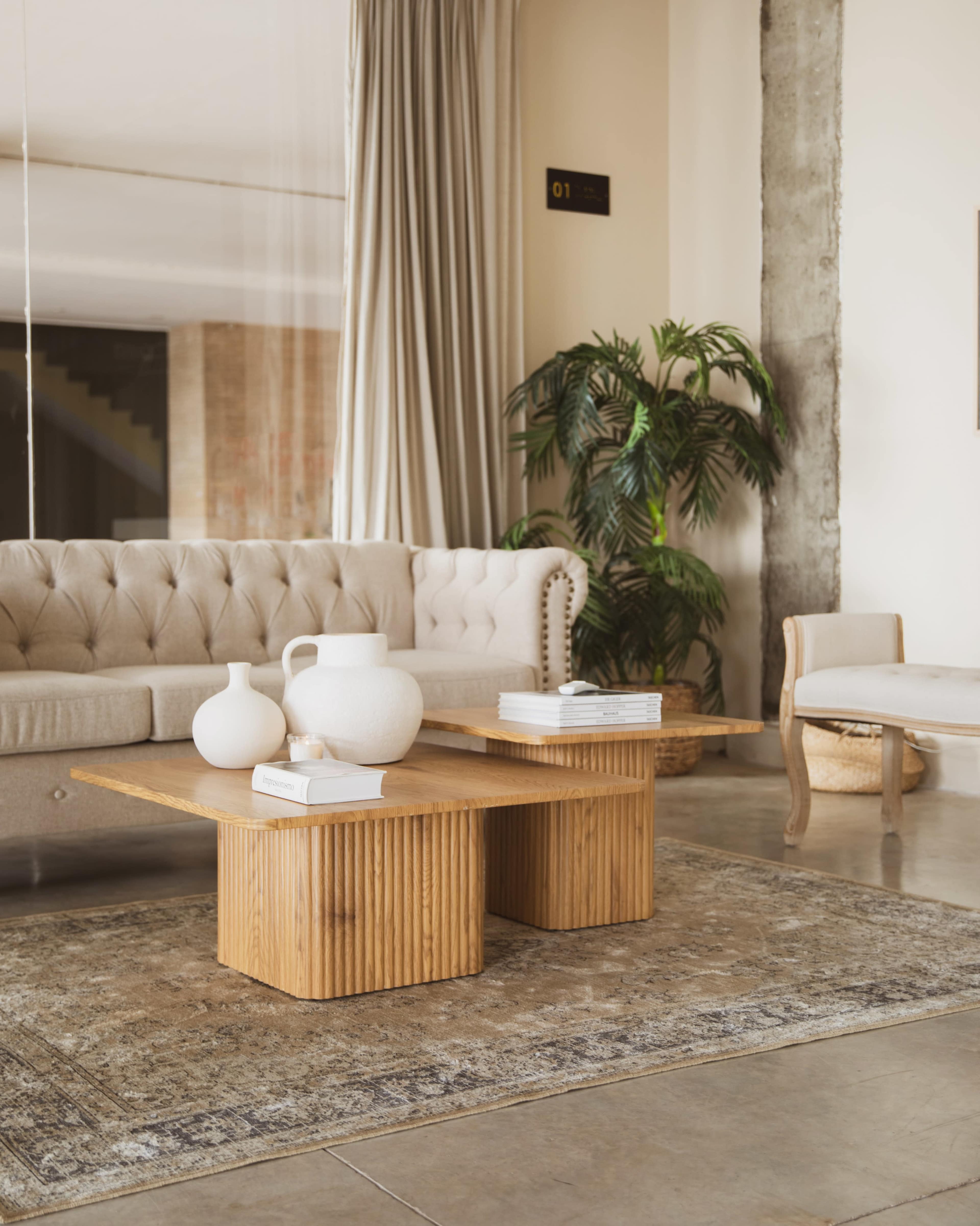 The image shows a modern living room featuring a light-colored tufted sofa, two wooden coffee tables, a potted plant, and a decorative rug.