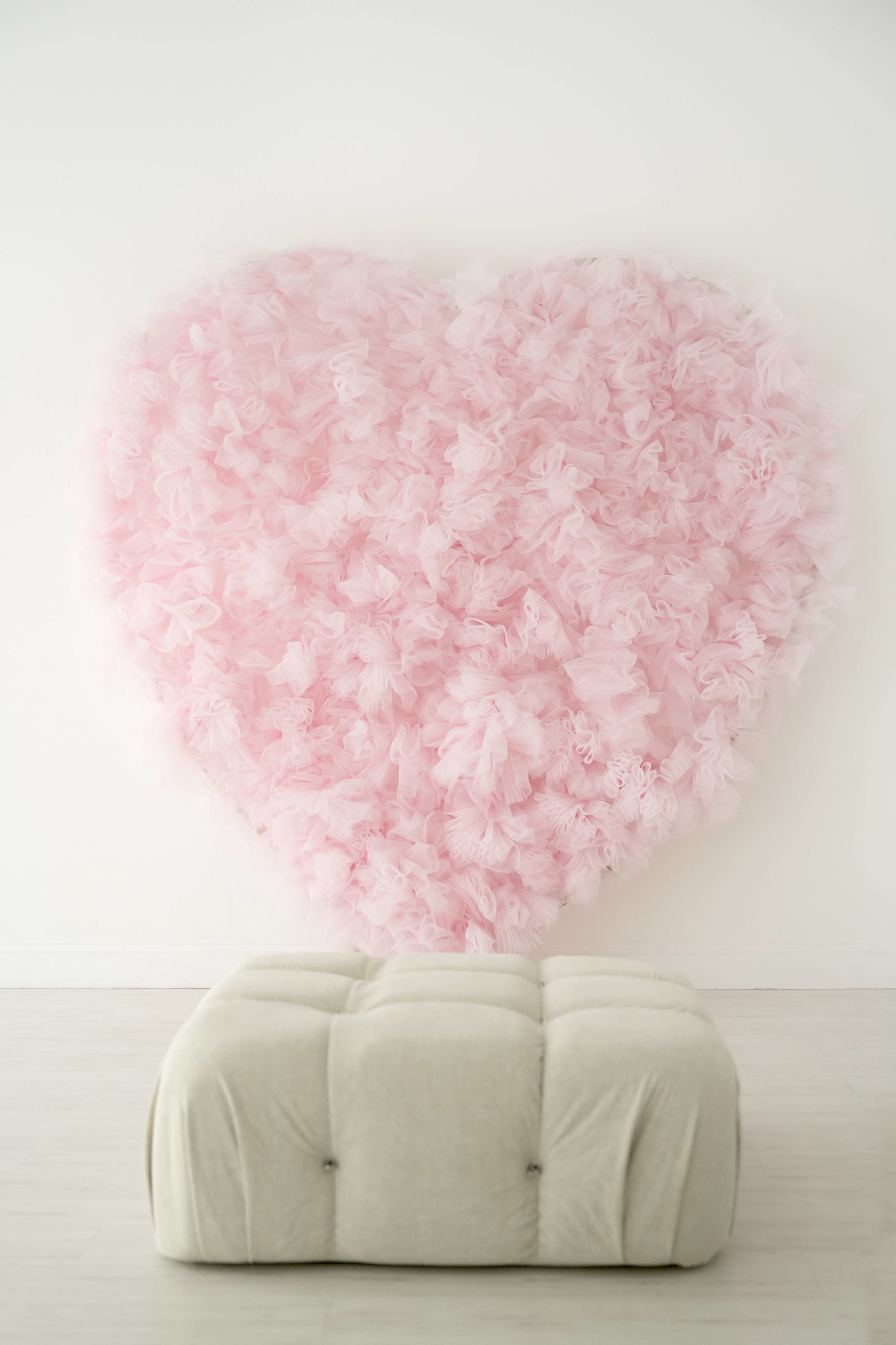 A large pink heart made of fluffy tulle fabric is mounted on a white wall above a beige tufted ottoman.