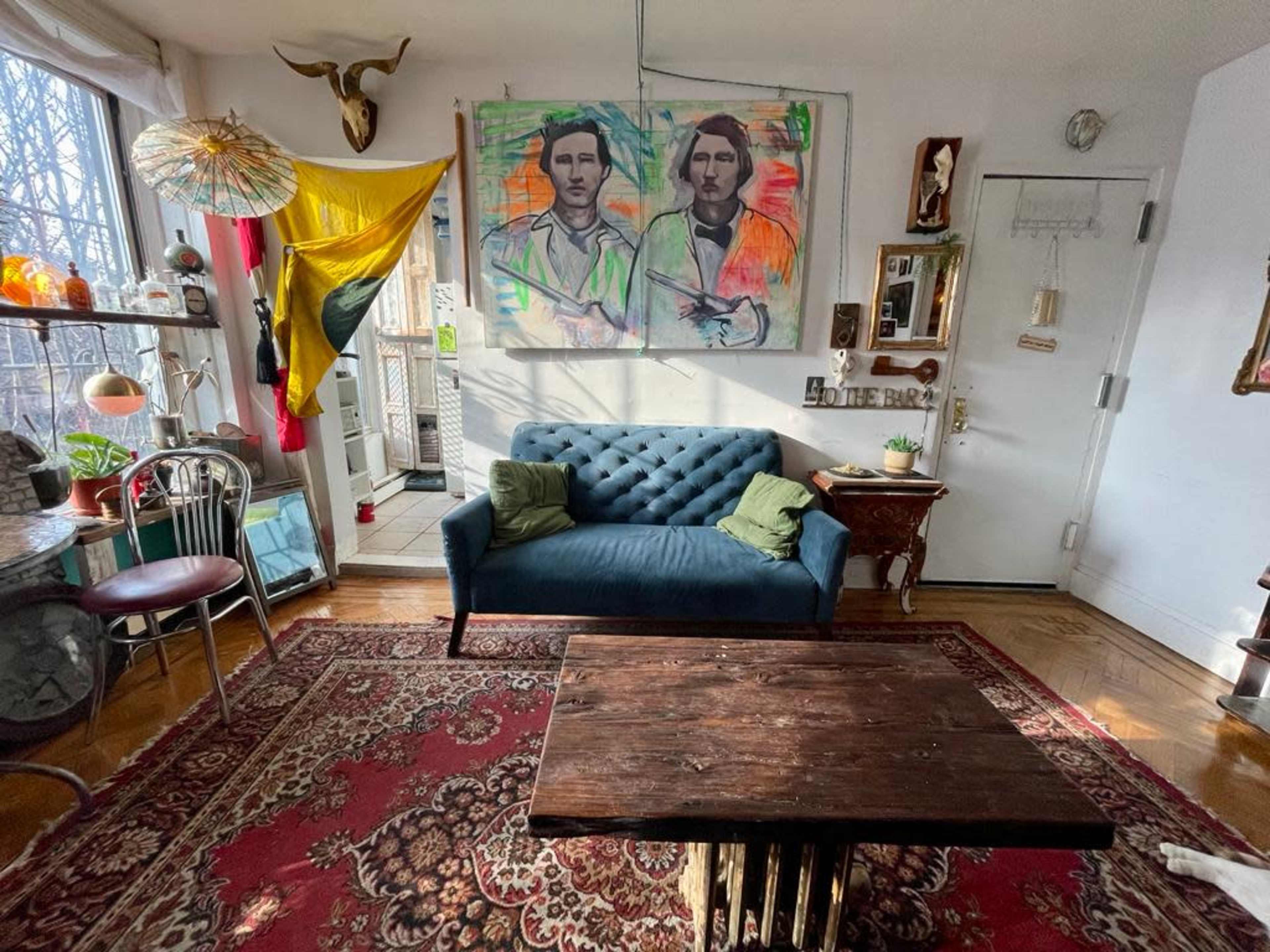 The image shows a living room with a blue velvet sofa, a wooden coffee table, and a large painting of two figures on the wall, accompanied by a yellow flag and various decorative items.