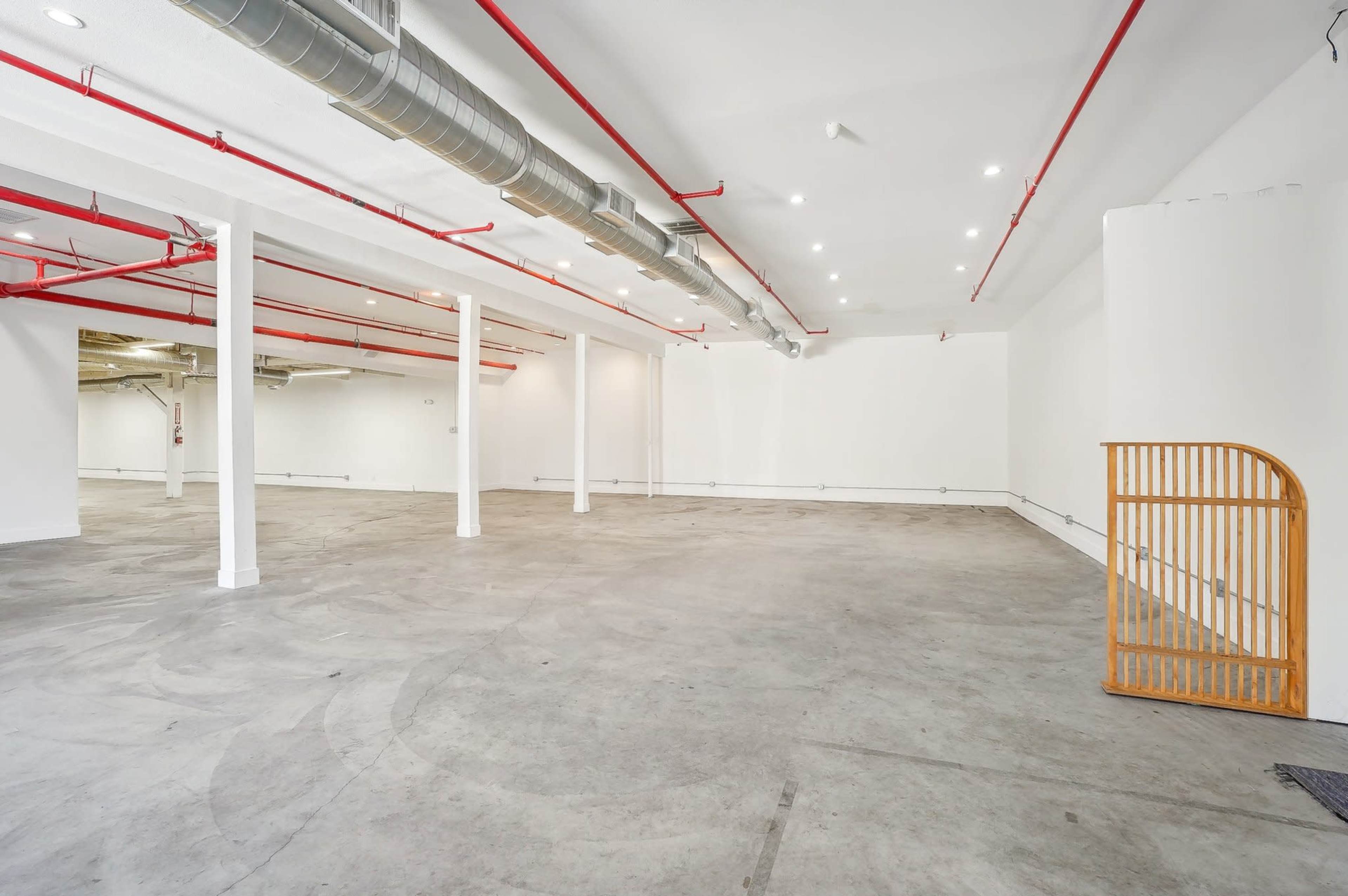The image shows a large, empty room with a concrete floor, white walls, red piping, and several overhead lights.