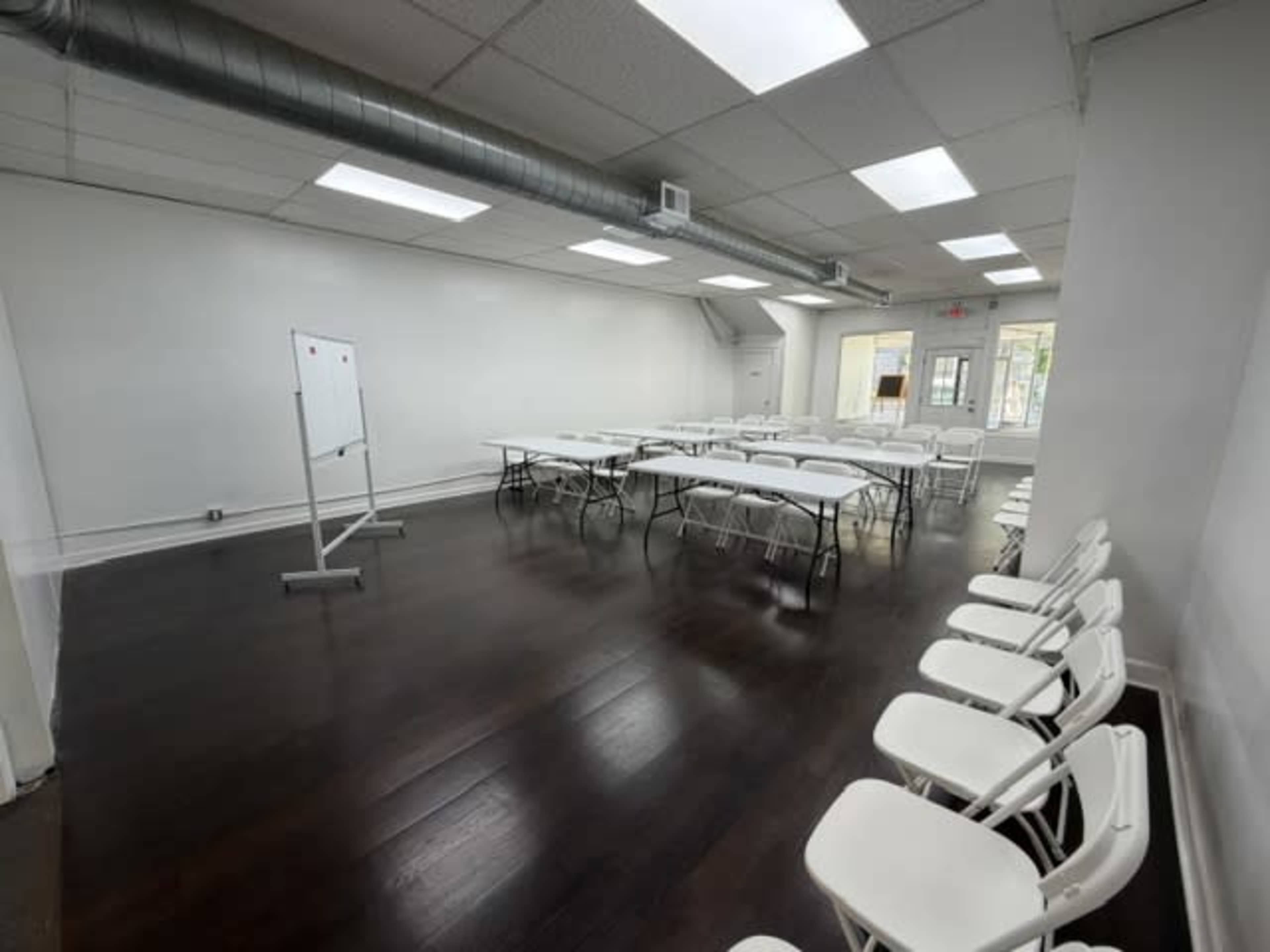The image shows a spacious, empty meeting room with white walls, rows of folding chairs, and a whiteboard on a stand.